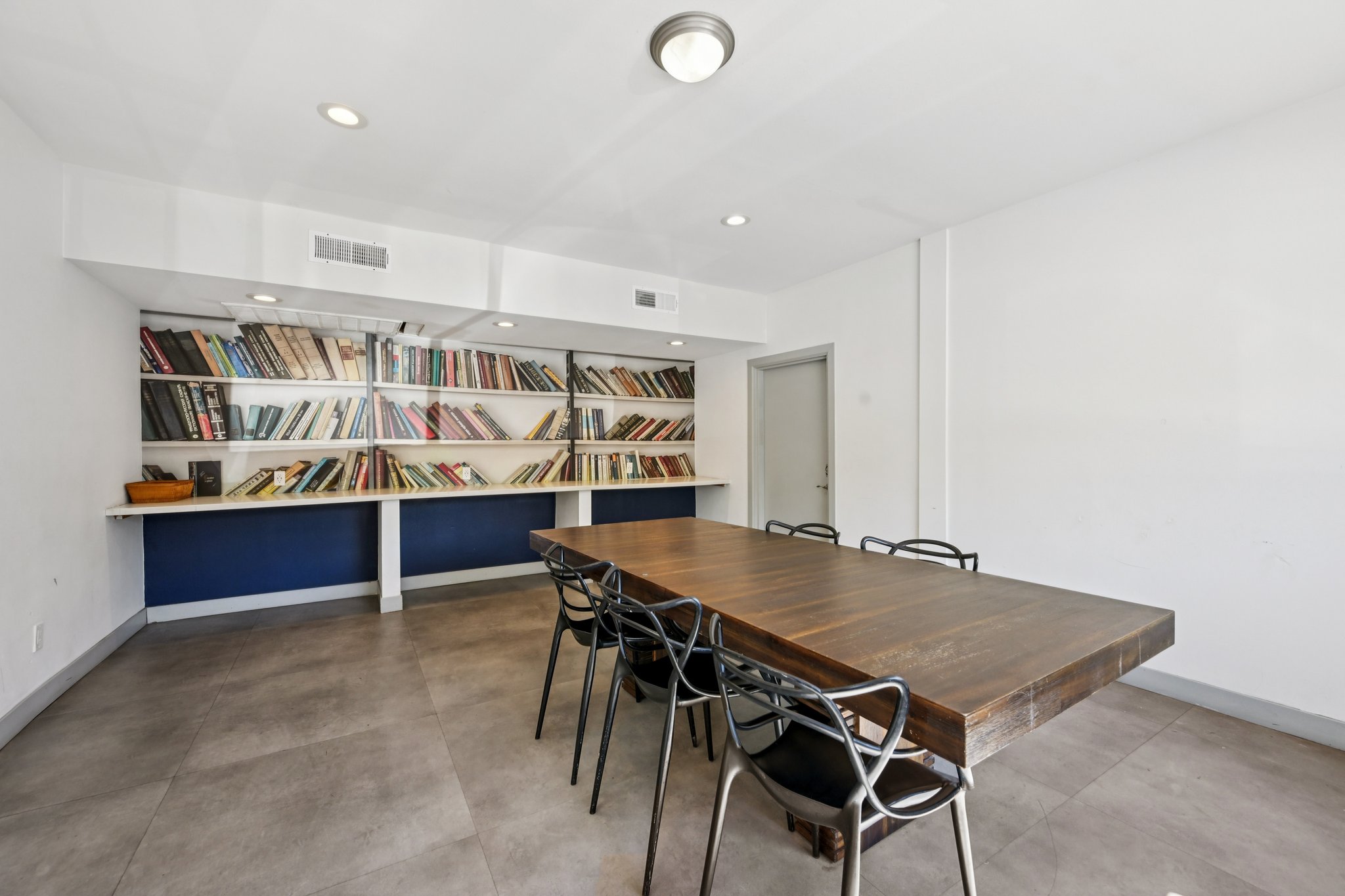 A spacious modern room featuring a large wooden dining table surrounded by black chairs. Behind the table is a stylish bookshelf filled with various books, and the walls are painted white with a blue accent section. The flooring is grey tiles, and there are recessed lights in the ceiling.