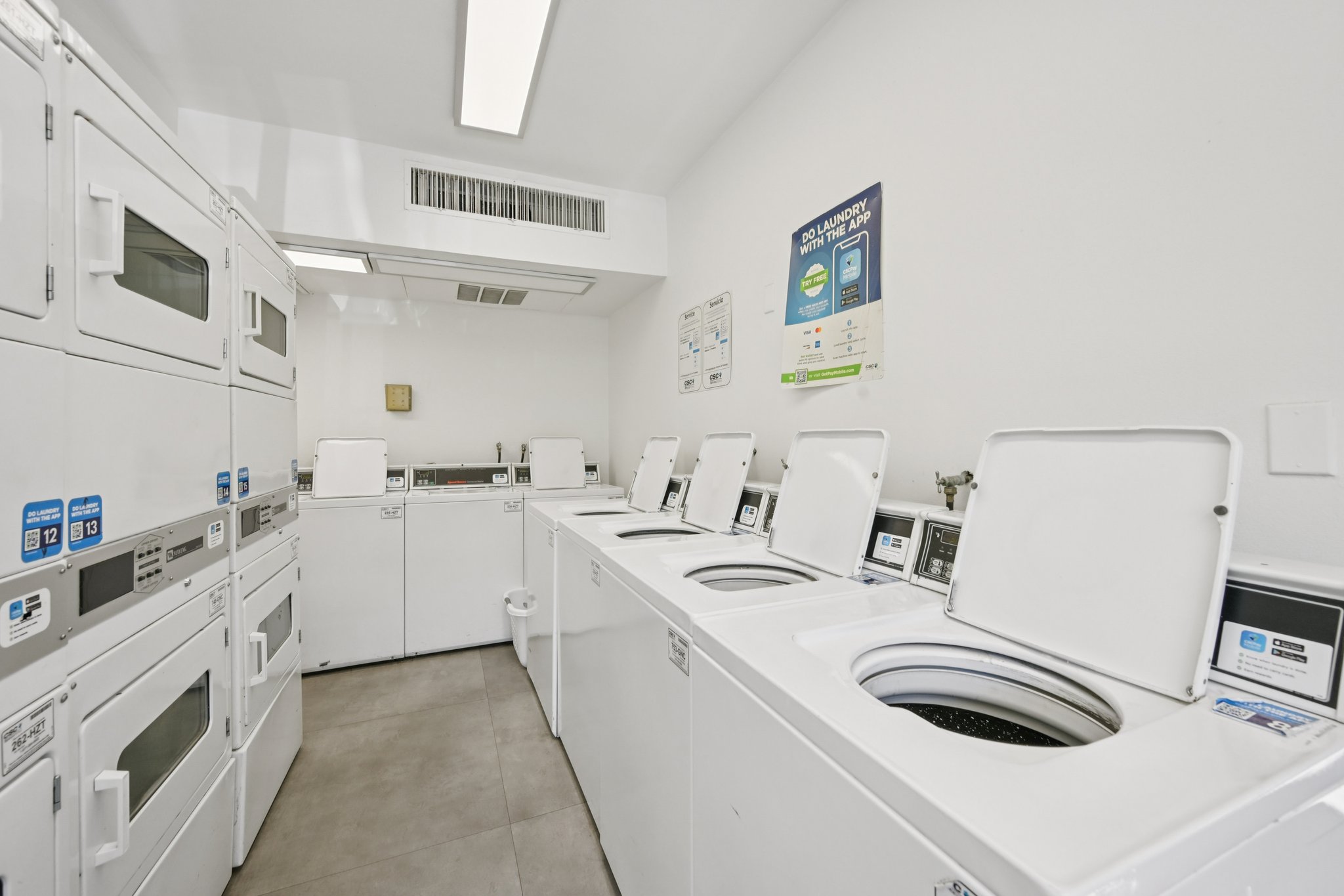 A clean, well-lit laundromat featuring several white washing machines and dryers lined up against a wall. Bright fluorescent lights illuminate the space, with informational signage visible on the wall. The floor is tiled with gray tiles, contributing to a modern and tidy appearance.