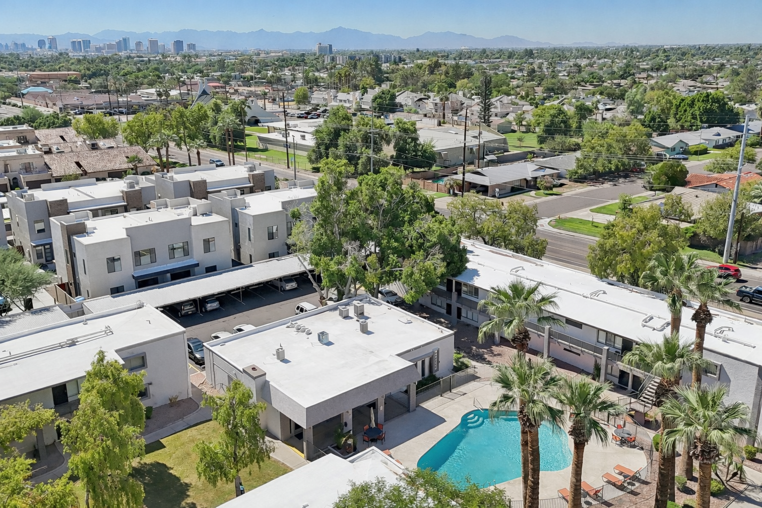 Aerial view of a residential area featuring low-rise buildings, a swimming pool surrounded by palm trees, and nearby green spaces. In the background, mountains and a clear blue sky add to the landscape, while various houses are visible across the neighborhood.