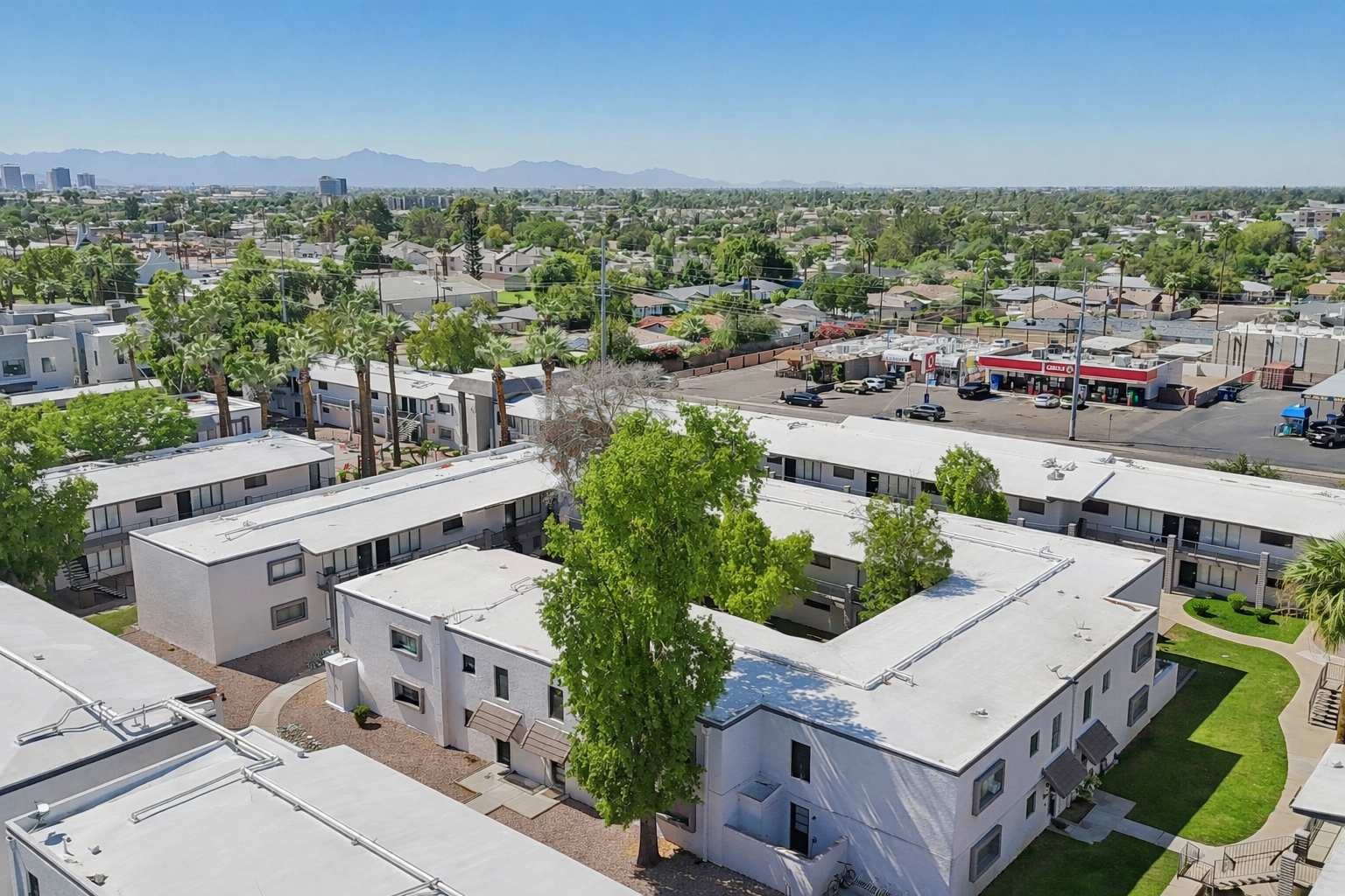 Aerial view of a residential complex with white-roofed buildings surrounded by green trees and landscaped areas. In the background, a cityscape can be seen, featuring low-rise buildings and mountains. The scene is bright and sunny, showcasing a suburban neighborhood.