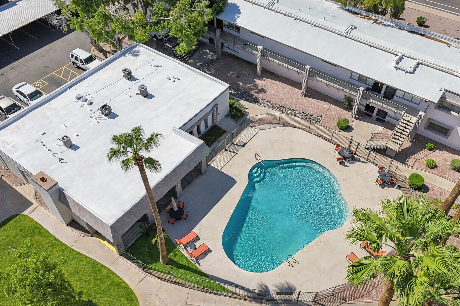 Aerial view of a modern hotel complex featuring a turquoise swimming pool surrounded by palm trees and lounge chairs. The building has a flat roof and is situated in a landscaped area with greenery and a paved pathway.