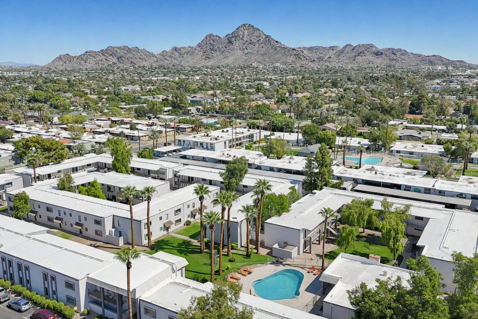 Aerial view of a residential area featuring low-rise apartment buildings surrounded by palm trees, with a swimming pool in the courtyard. In the background, a mountain range rises against a clear blue sky, showcasing the natural landscape. The scene captures a blend of urban living and nature.