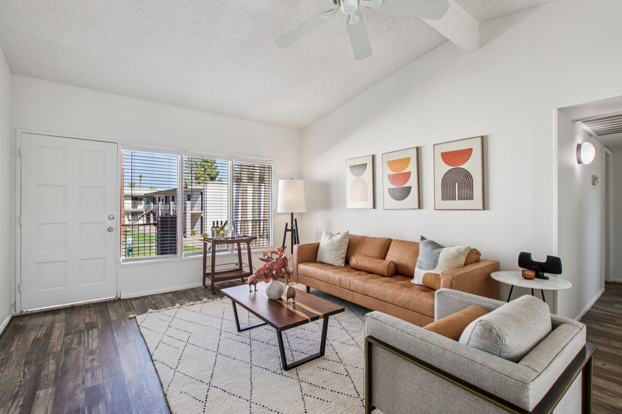 Modern living room featuring a brown leather sofa, a light gray armchair, and a wooden coffee table. A patterned area rug is on the floor, and artwork hangs on the white walls. Large windows allow natural light to flood the space, with a view of the outdoors. Ceiling fan and floor lamp add to the decor.