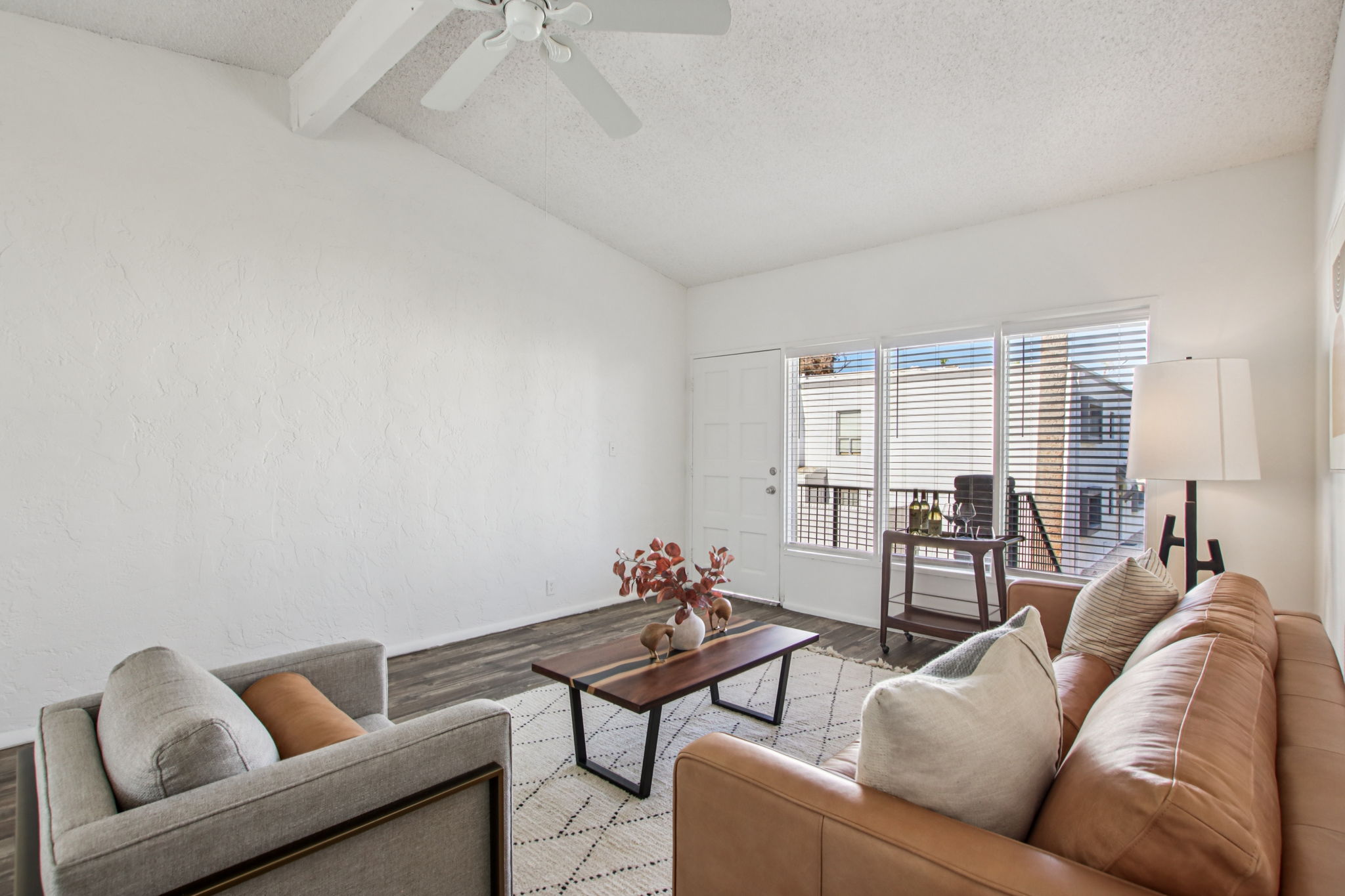 A bright and spacious living room featuring a leather sofa, a light gray armchair, and a coffee table on a patterned rug. Large windows provide ample natural light, with a potted plant and a small desk visible in the background. The walls are white, and the ceiling is high with a ceiling fan.