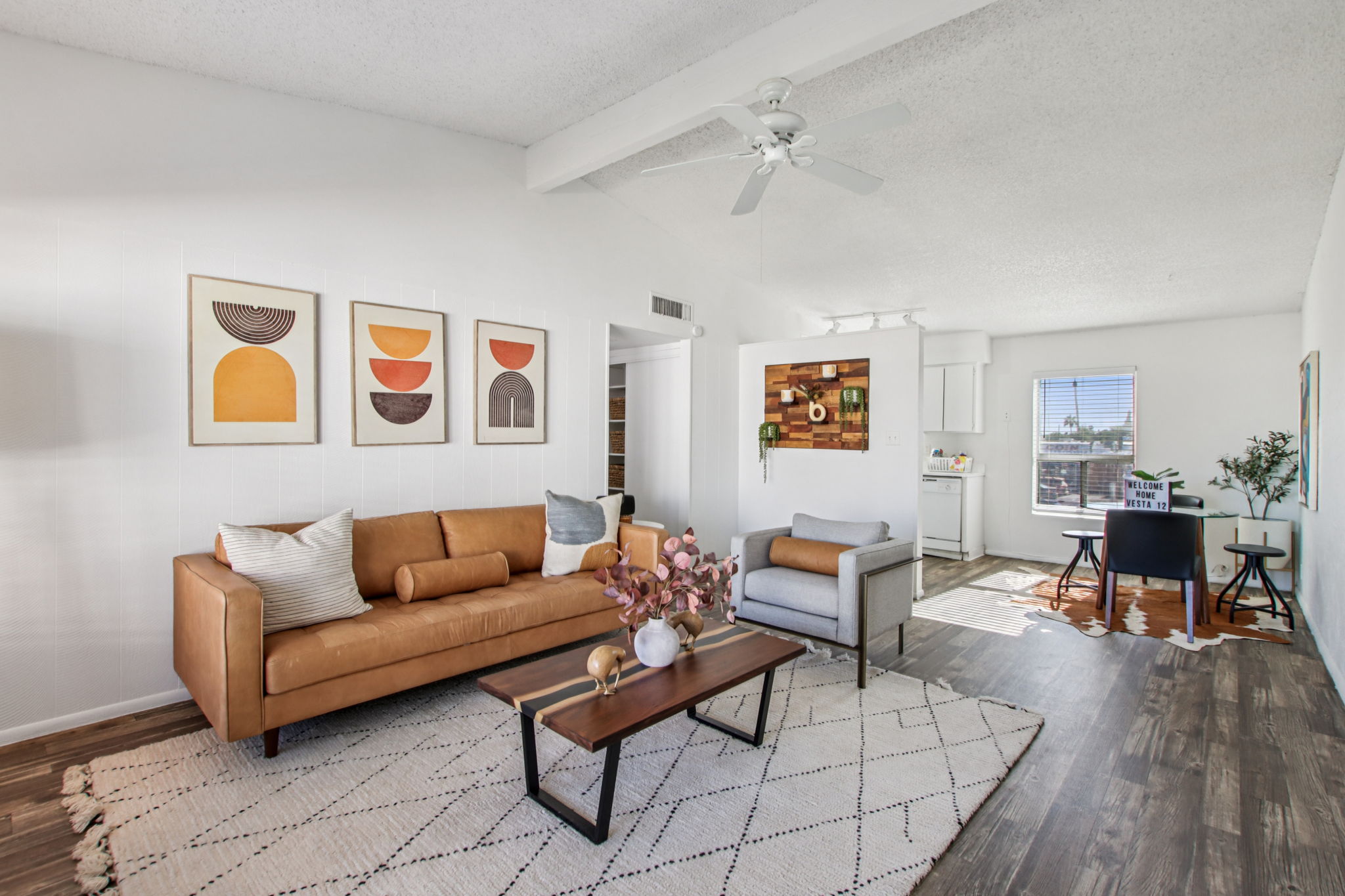 A modern living room featuring a brown leather sofa with decorative pillows, a wooden coffee table with a vase of pink flowers, and a cozy armchair. Wall art in various shapes and colors adorns the walls. Natural light floods in from a window, highlighting a stylish, contemporary aesthetic.
