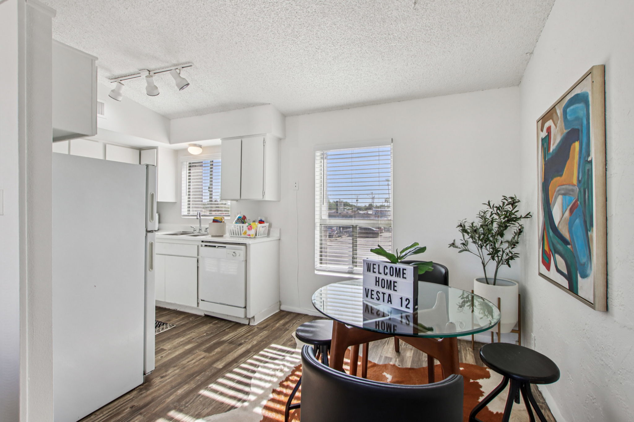 A bright, modern kitchen and dining area featuring a white refrigerator, stove, and dishwasher. A round glass dining table with black chairs sits near a window, alongside a decorative plant. A "Welcome Home" sign is displayed on the table, and a colorful abstract painting adorns the wall. Natural light streams in through the window.