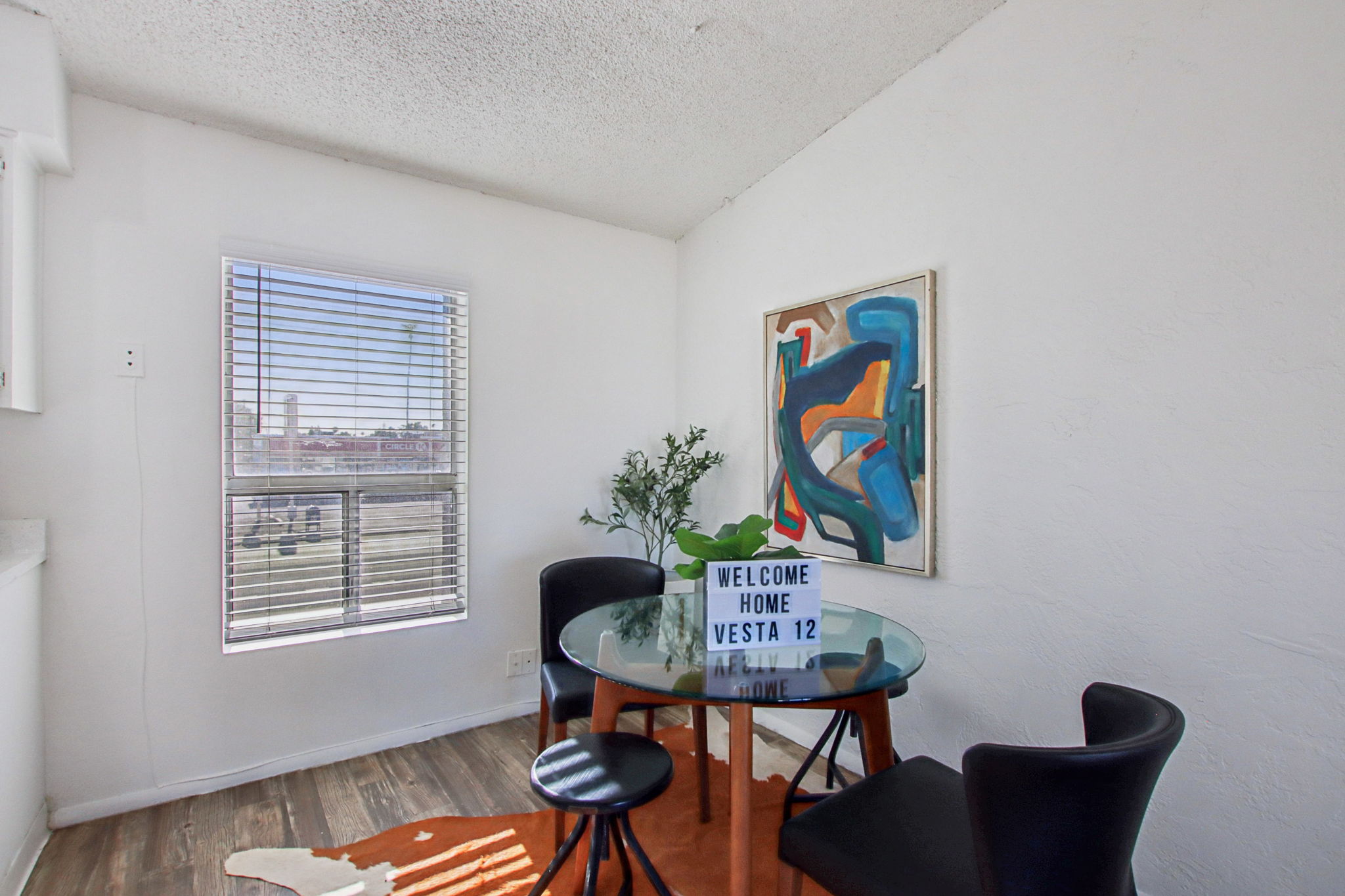 A bright and modern dining area featuring a round glass table with three black chairs. A decorative plant sits on the table next to a sign reading "WELCOME HOME VESTA 12." The walls are white, and there's an abstract painting on display. A window provides natural light, showcasing a view outside.