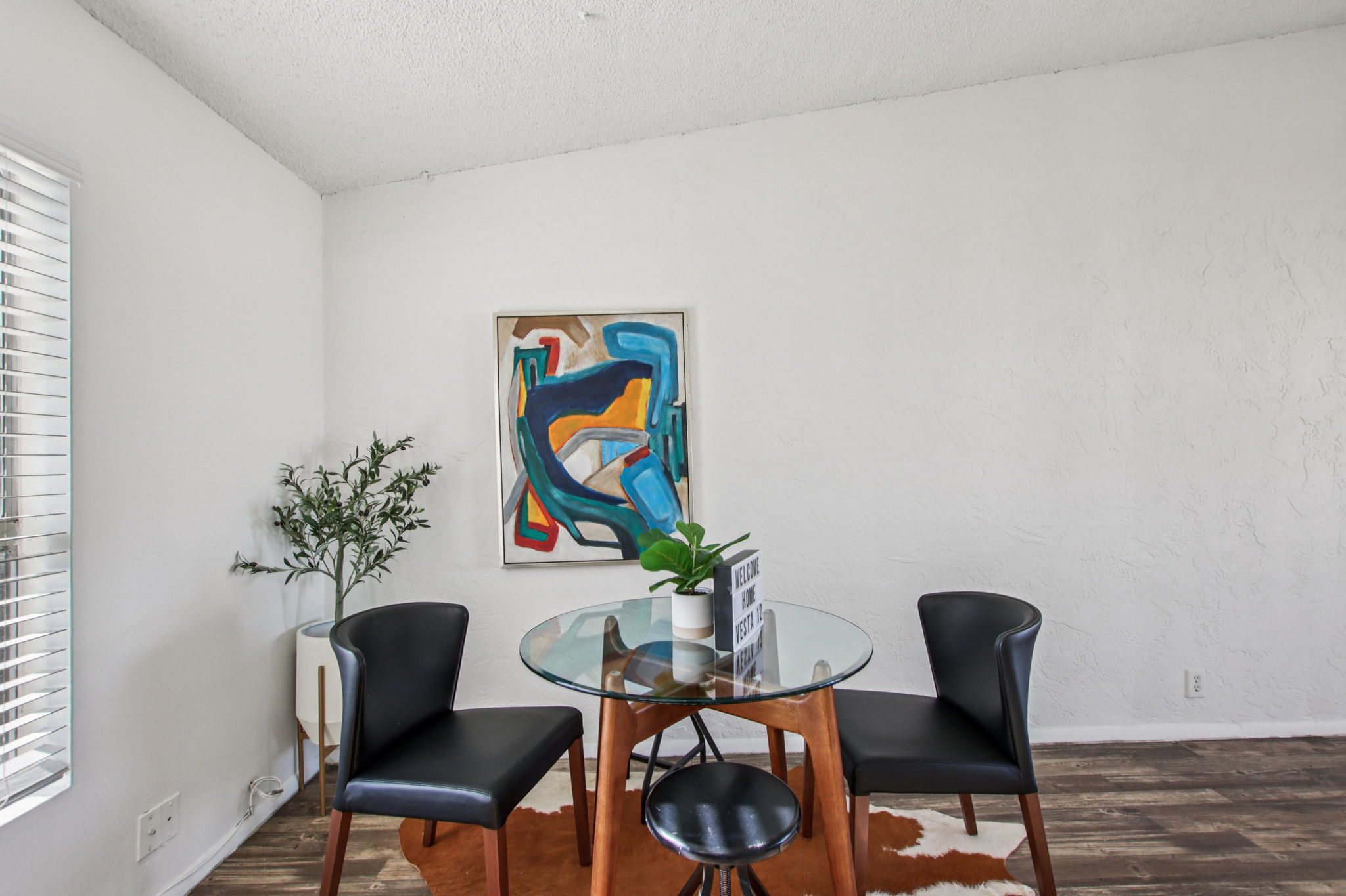 A bright, modern dining area featuring a round glass table with two black chairs. There's an abstract painting on the wall and a small plant on the table. The flooring is wood, and there are large windows, allowing natural light to fill the space. The walls are painted white, creating a clean, open atmosphere.