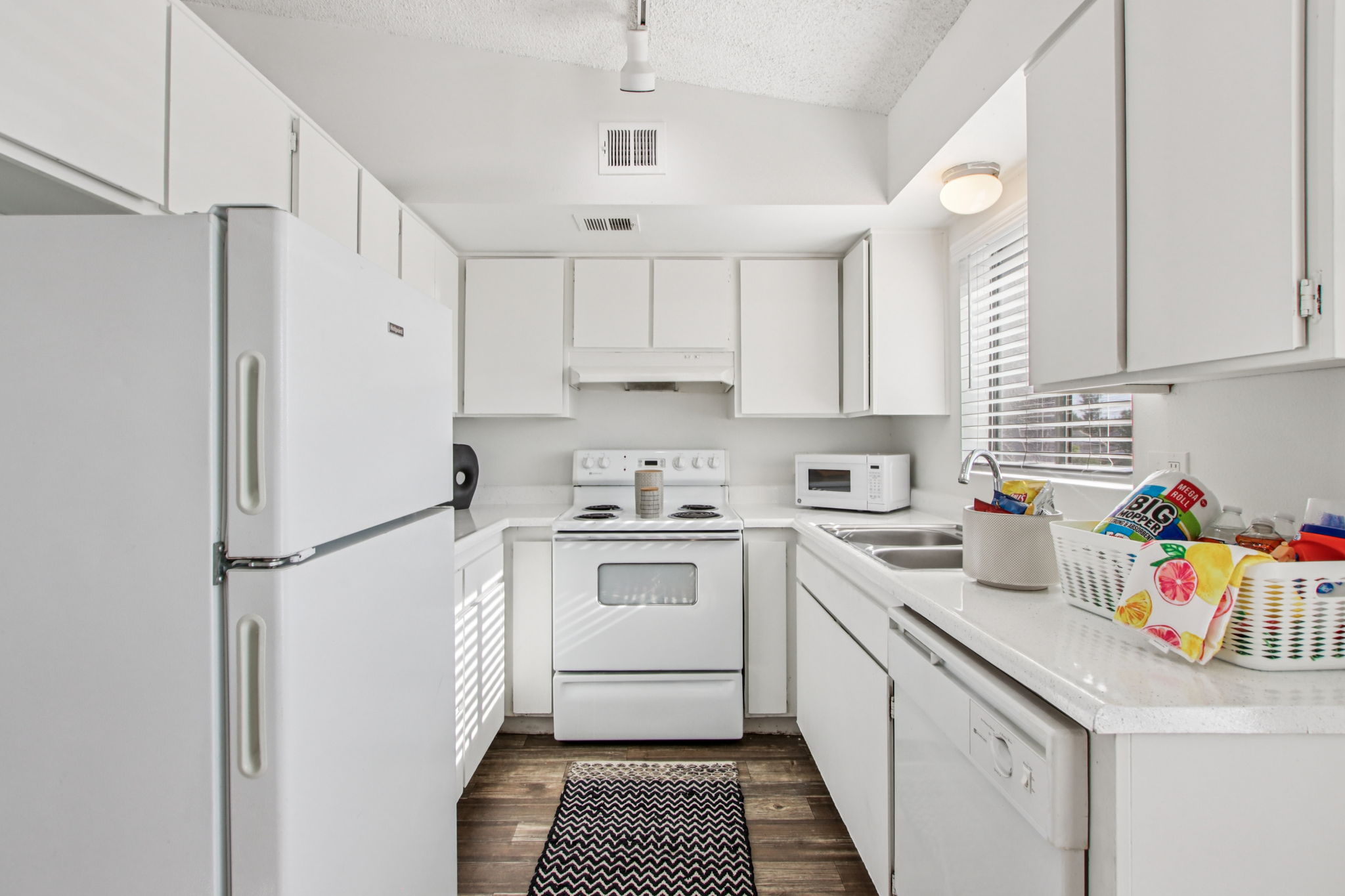 Bright and modern kitchen featuring white cabinets and appliances, a refrigerator, stove, and microwave. The countertop is well-organized, with a basket of colorful items. Natural light streams in through a window, highlighting the space's clean and open layout.