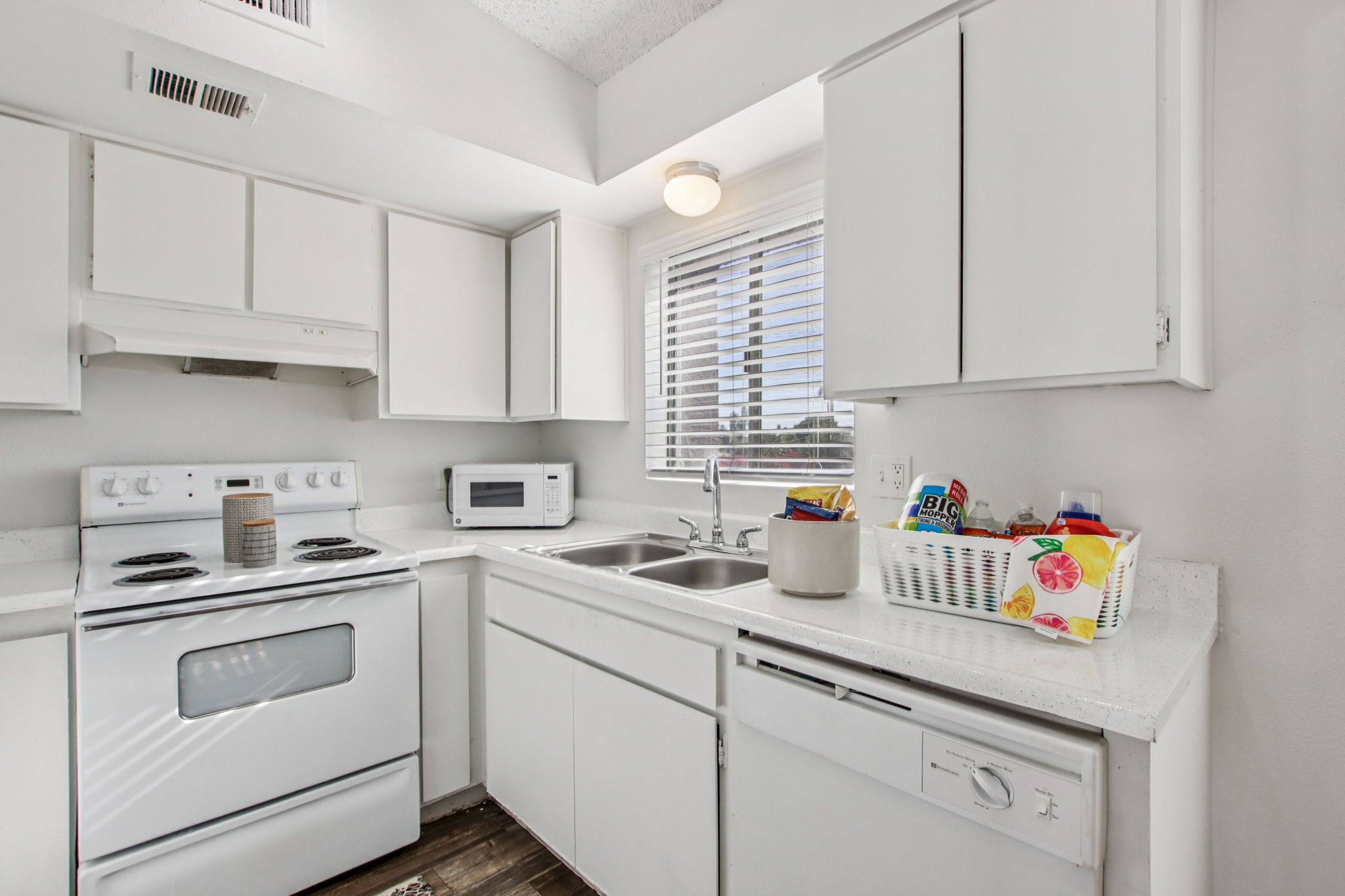 A bright, modern kitchen featuring white cabinets, a stove, an oven, a microwave, and a double sink. Natural light streams through a window with blinds, illuminating the countertop, which holds a basket filled with colorful kitchen items. The floor is wooden, adding warmth to the space.