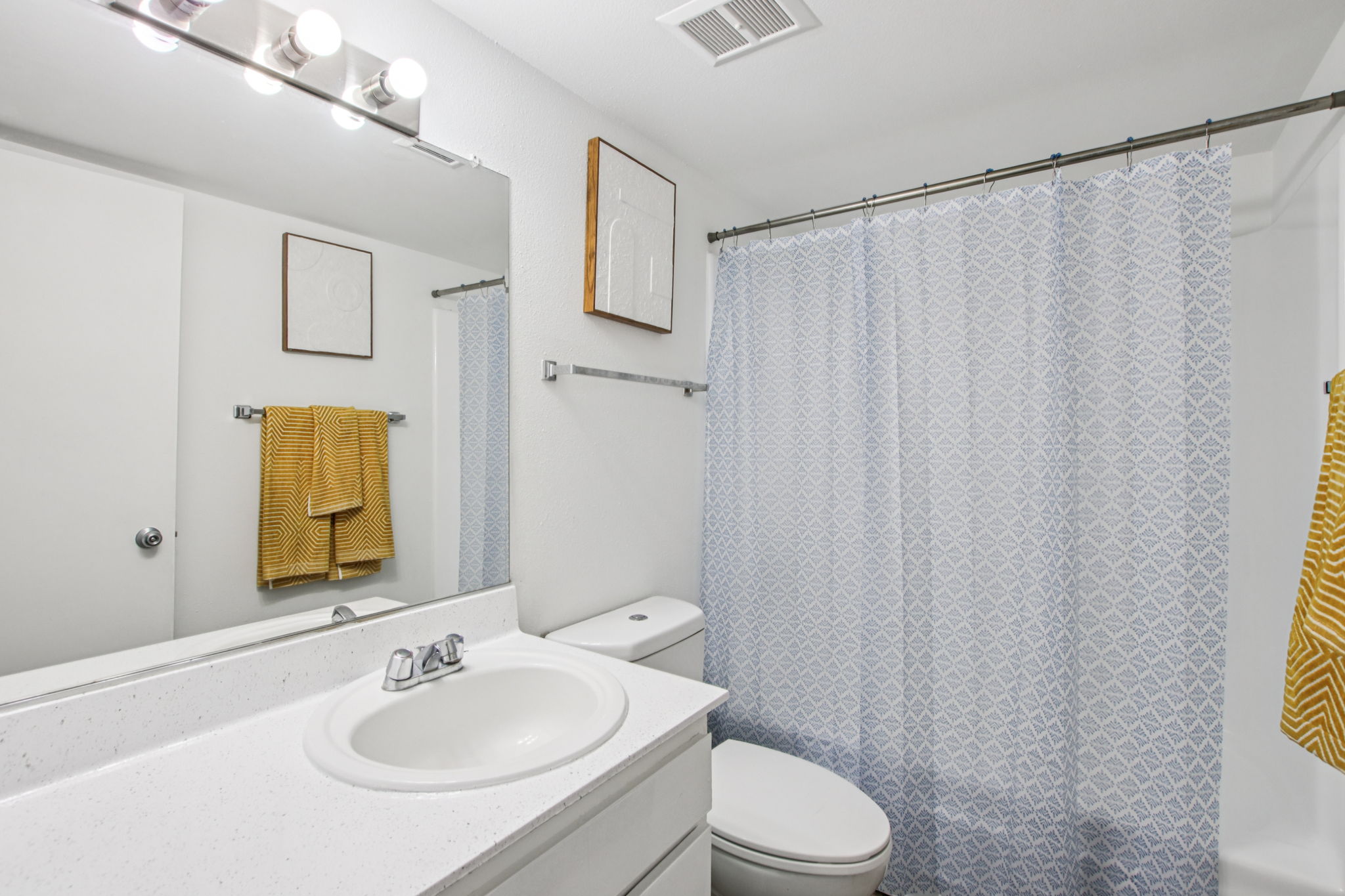 A clean, bright bathroom featuring a white countertop with a sink, a mirror above, and a shower curtain with a light blue geometric pattern. There are two yellow towels hanging on a rack and a toilet in the corner. The walls are painted white, and there is a small piece of art on the wall.