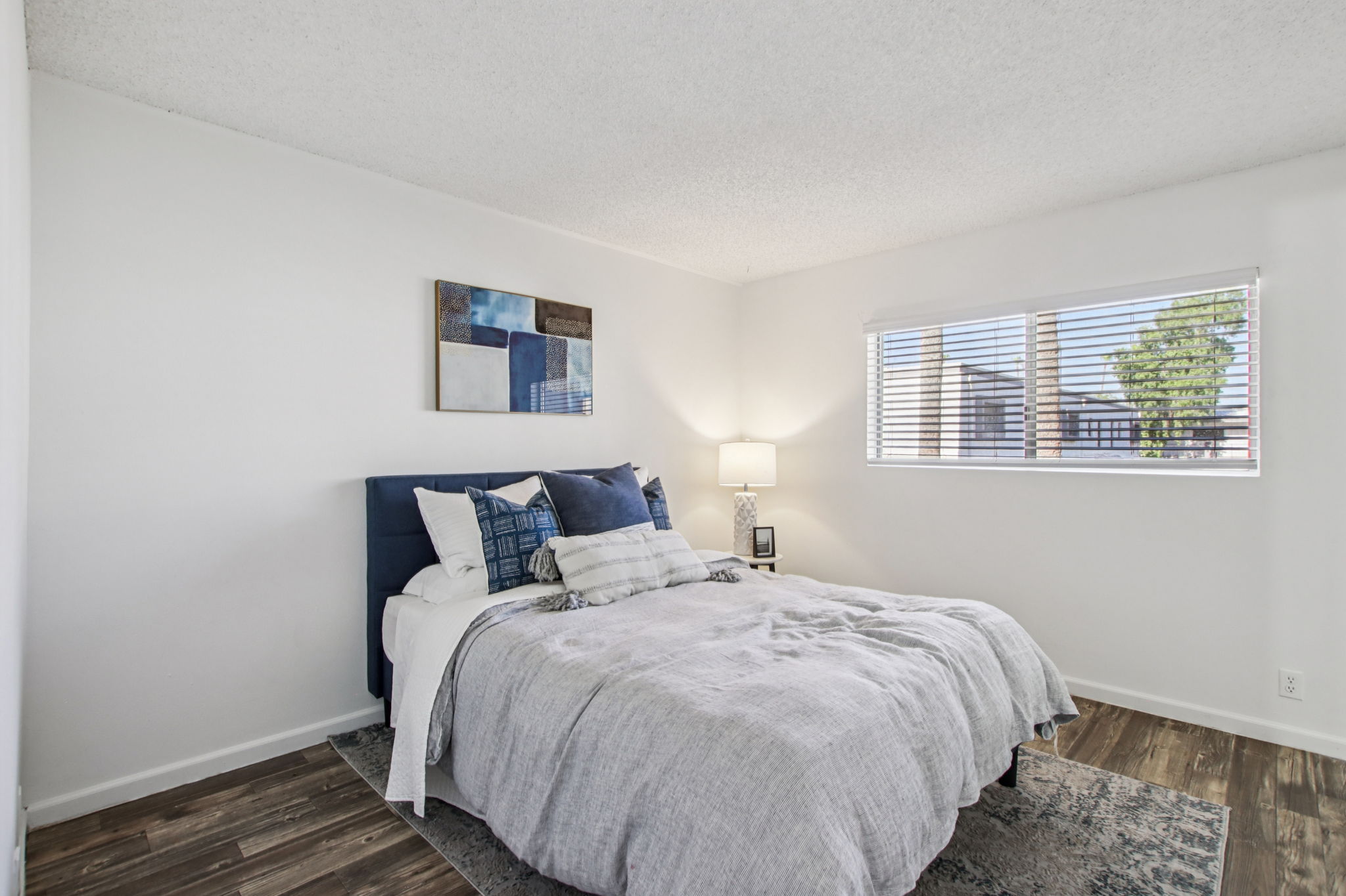 A cozy bedroom featuring a neatly made bed with blue and white bedding. A bedside table with a lamp sits next to the bed. A window lets in natural light, revealing a view of trees outside. The walls are painted white, and the floor is a warm wooden laminate, adding to the inviting atmosphere.