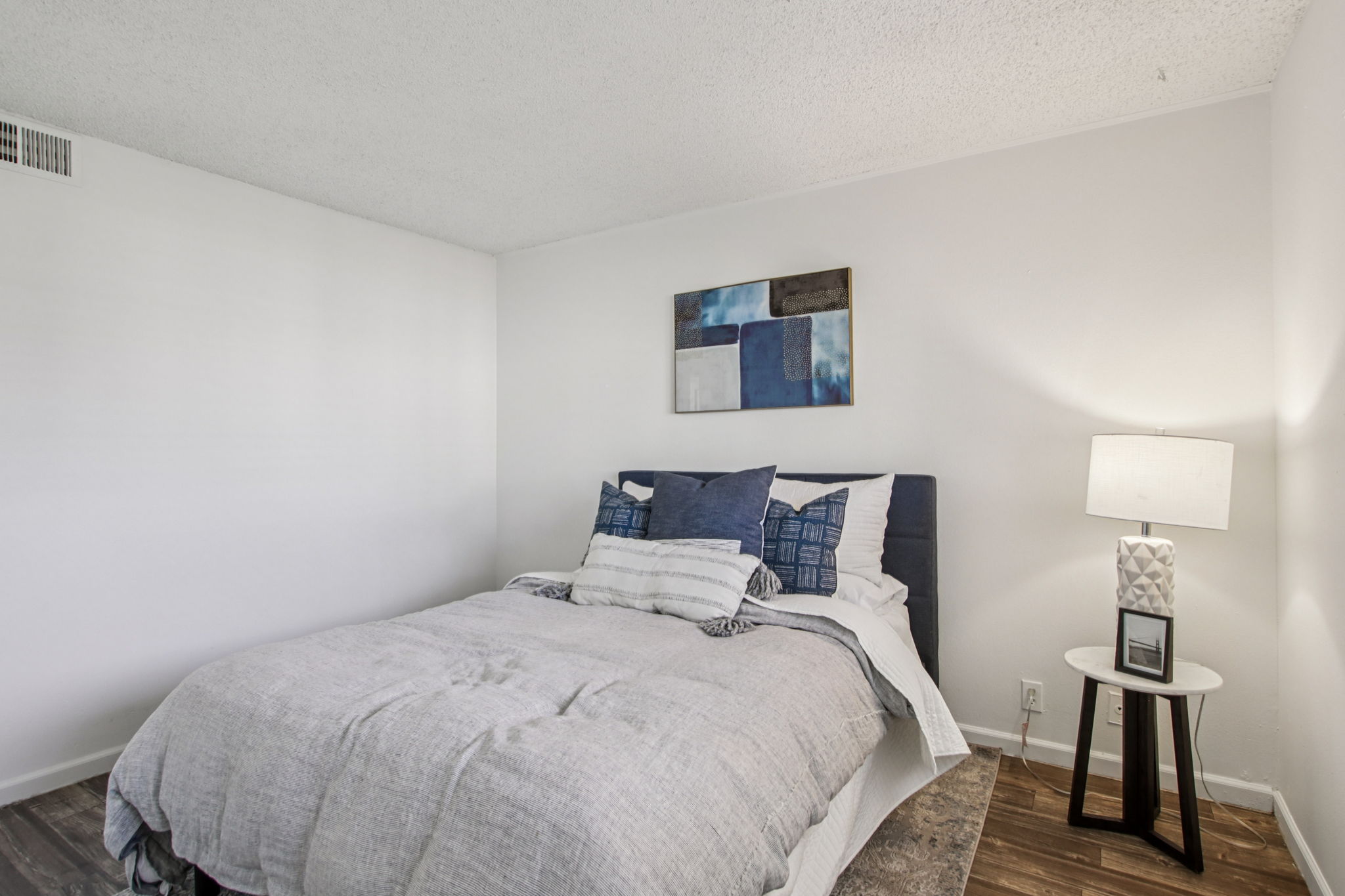 A cozy bedroom featuring a double bed with gray bedding and assorted blue throw pillows. A small side table with a white lamp is beside the bed, and a modern abstract painting hangs on the wall above the bed. The room has light-colored walls and a wooden floor, creating a warm, inviting atmosphere.