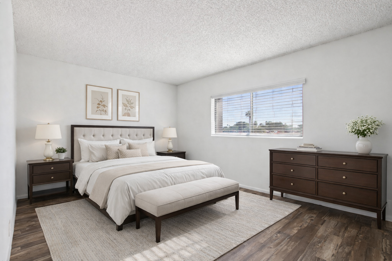 A modern bedroom featuring a queen-sized bed with neutral-colored bedding and decorative pillows. Flanking the bed are two nightstands with lamps. A large window lets in natural light, and there's a wooden dresser and a bench at the foot of the bed. The room has a cozy, inviting atmosphere with a light color palette.