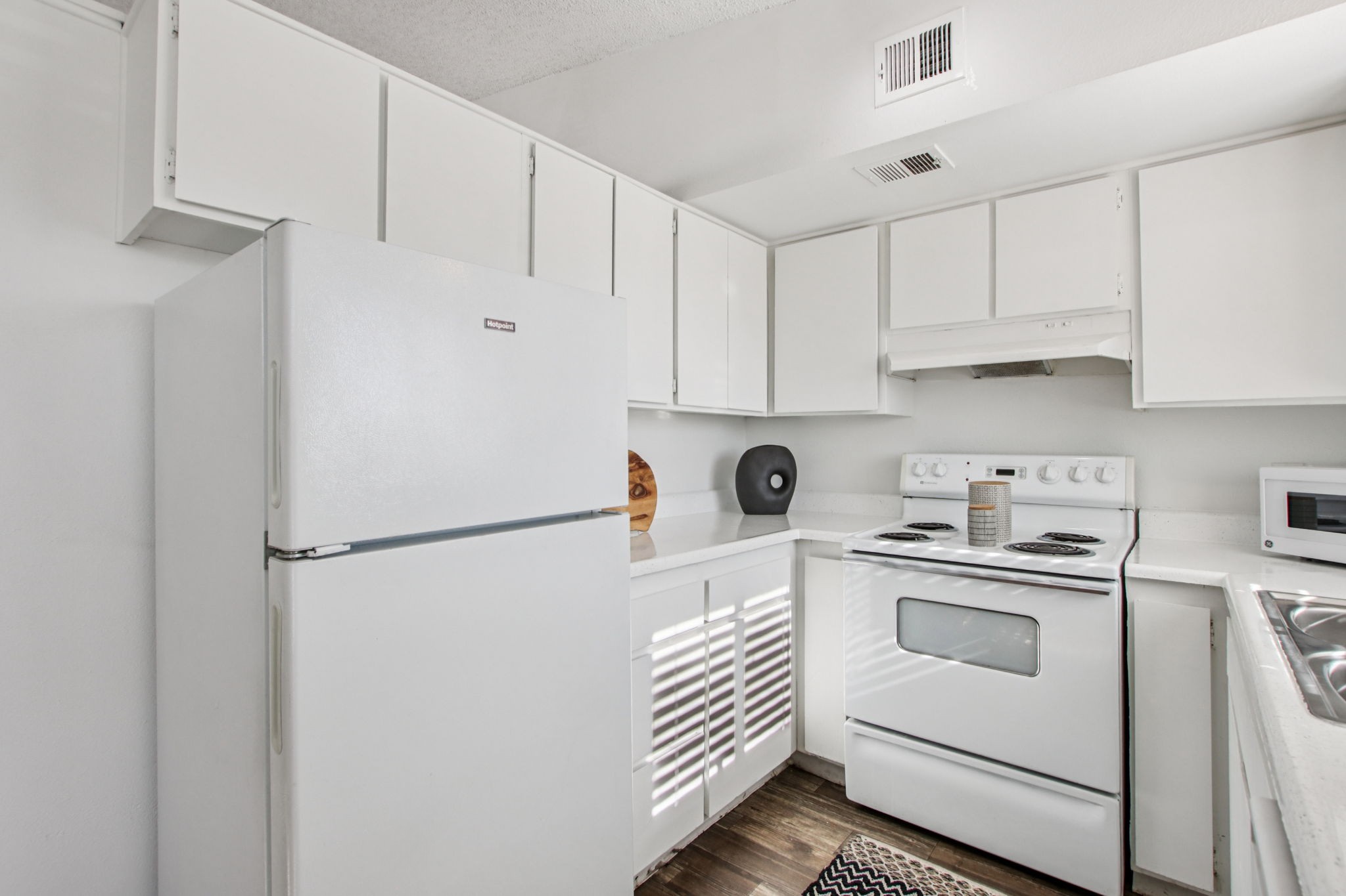 Bright, modern kitchen featuring white cabinetry, a white refrigerator, an electric stove, and a microwave. The countertop is clean and minimalistic, with a black decorative item and a patterned rug on the floor. Natural light filters through the space, creating a fresh and airy atmosphere.