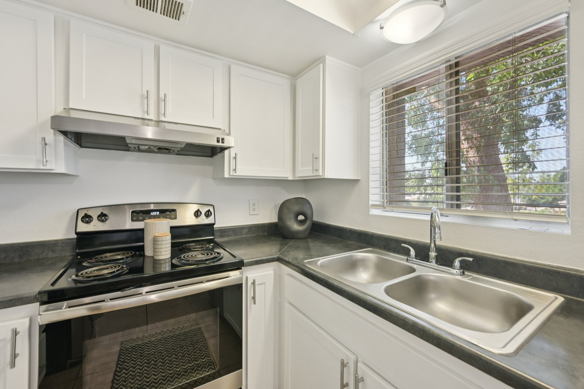 A modern kitchen featuring a stainless steel stove with four burners, a double sink, and white cabinetry. A window with blinds allows natural light to enter, showcasing greenery outside. The countertop is dark, contrasting with the white cabinets, creating a clean and contemporary look.