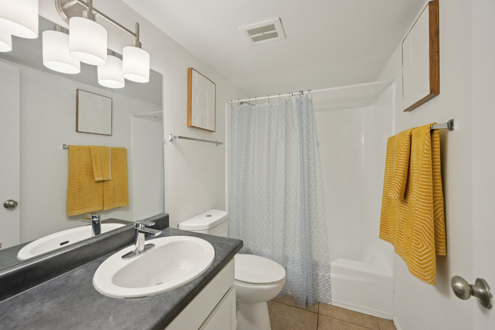 A bright and clean bathroom featuring a dark countertop with a sink, a modern mirror, and two yellow towels hanging on the wall. A light blue shower curtain separates the shower area, and there is a white toilet next to a tub. The walls are painted white, creating a fresh, airy atmosphere.