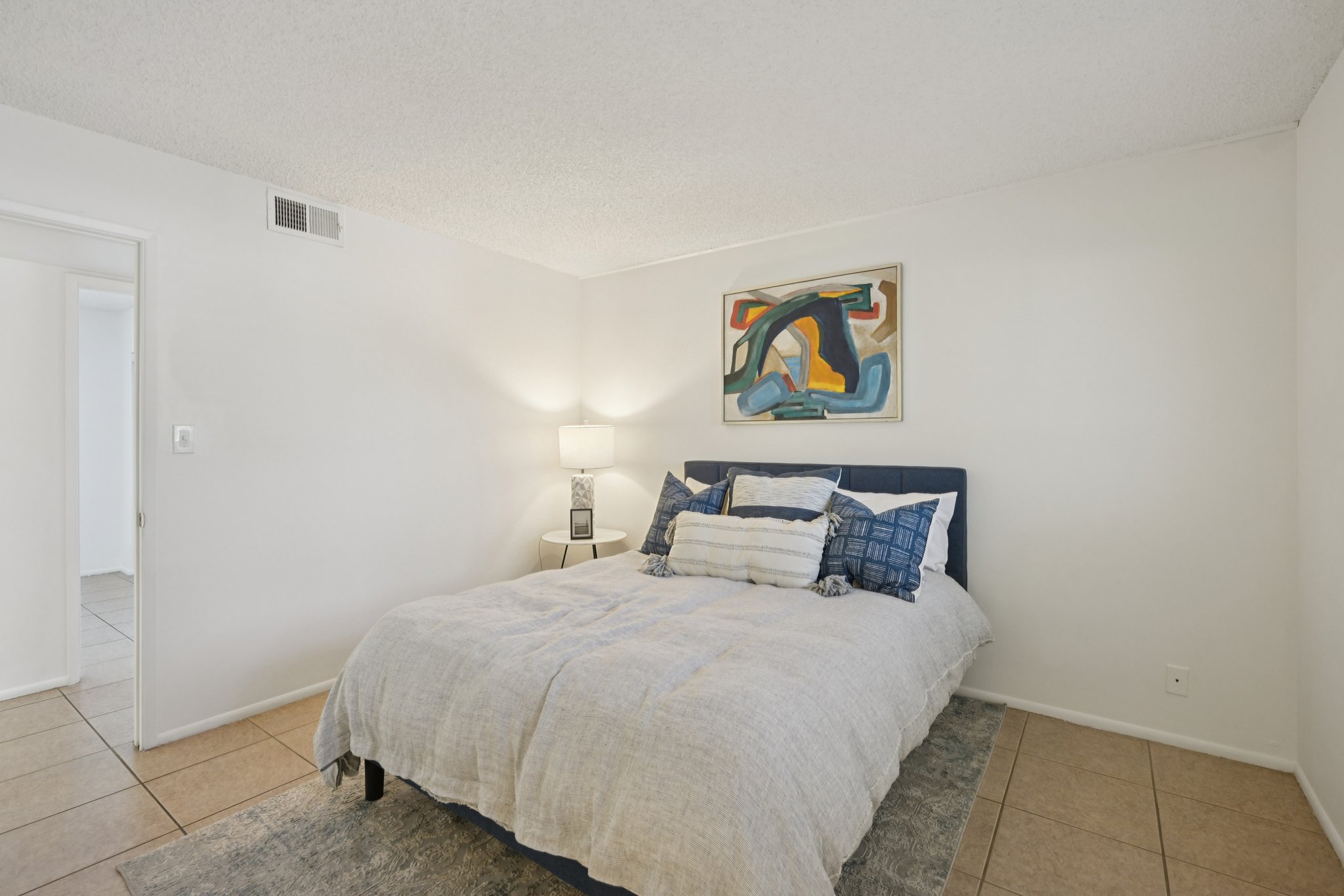 A cozy bedroom featuring a neatly made bed with blue and white bedding, a modern abstract painting on the wall above the bed, a bedside lamp on a small table, and a light-colored rug on tiled flooring. The room has neutral walls and a door leading to another space.