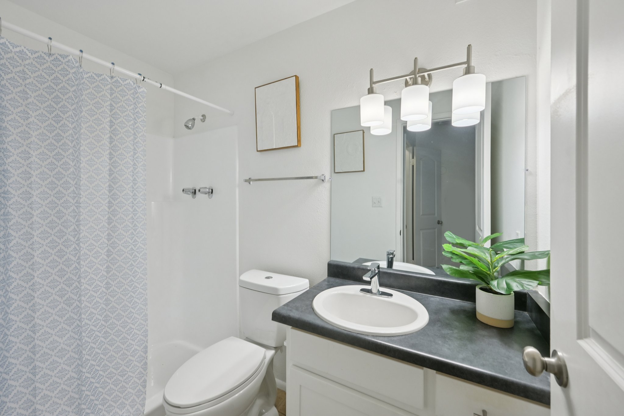 A modern bathroom featuring a white shower curtain with a blue geometric pattern, a sleek sink with a black countertop, a mirror with three light fixtures above, and a potted plant. The toilet is visible, and there is a small piece of artwork on the wall. The overall color scheme is neutral and bright.