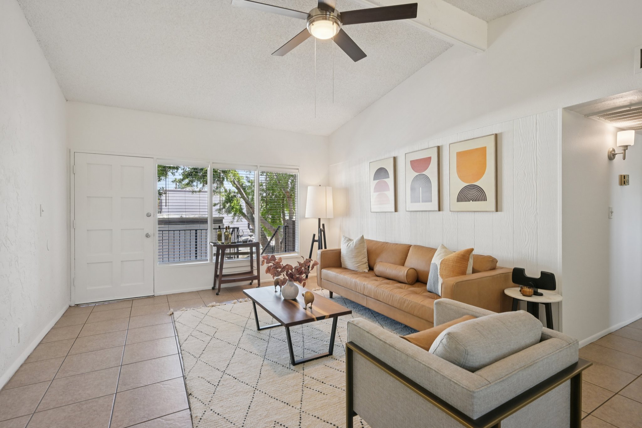 A modern, well-lit living room with a beige sofa and a gray armchair. There’s a coffee table in front of the sofa with decorative items. A large window allows natural light to enter, and framed artwork is displayed on the wall. The room features a ceiling fan and light-colored tiles on the floor.
