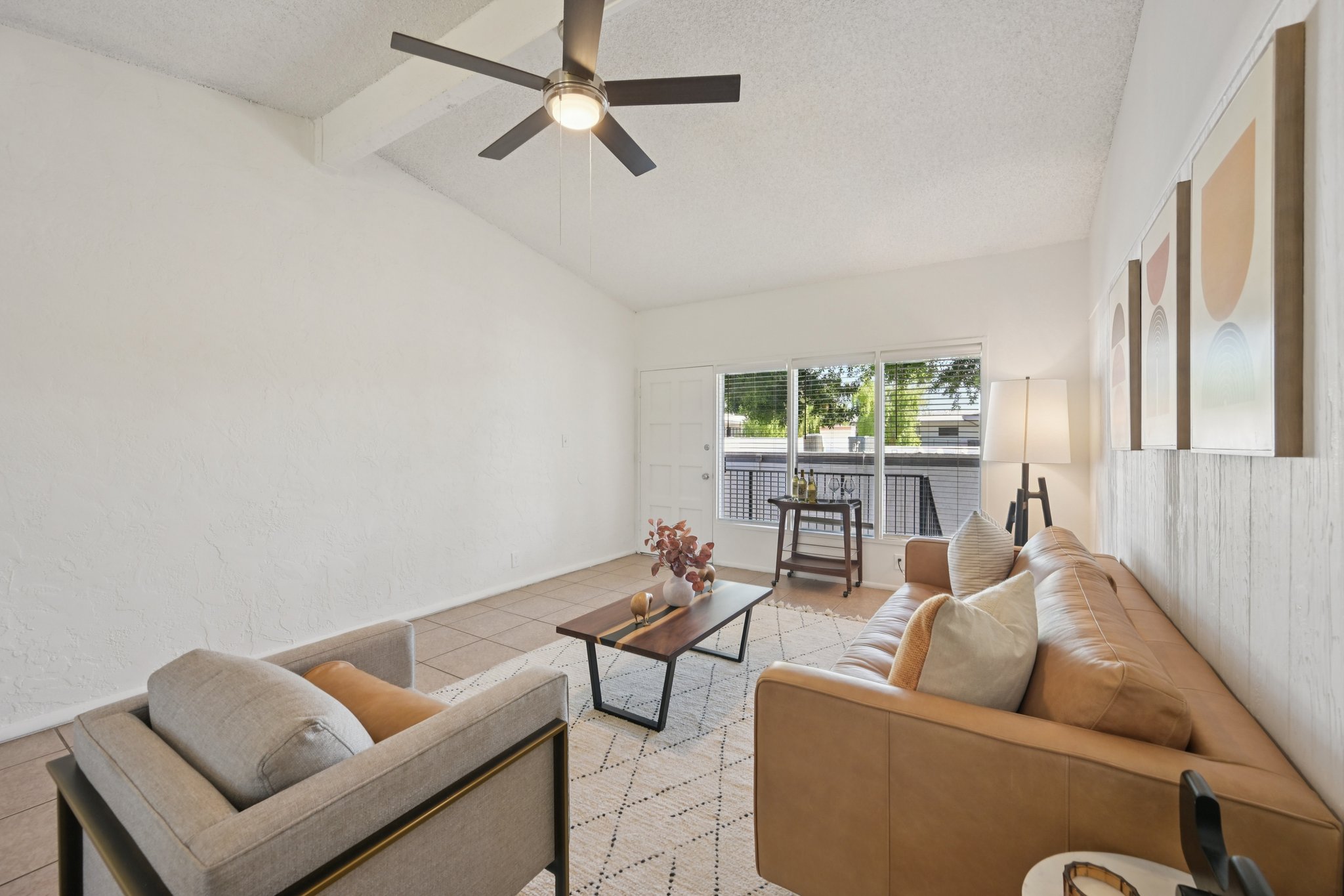 A bright and airy living room featuring a tan leather sofa, a light gray armchair, and a wooden coffee table. A ceiling fan hangs from the textured ceiling, and large windows allow natural light to fill the space. Decorative wall art and a floor lamp enhance the modern, minimalist aesthetic.