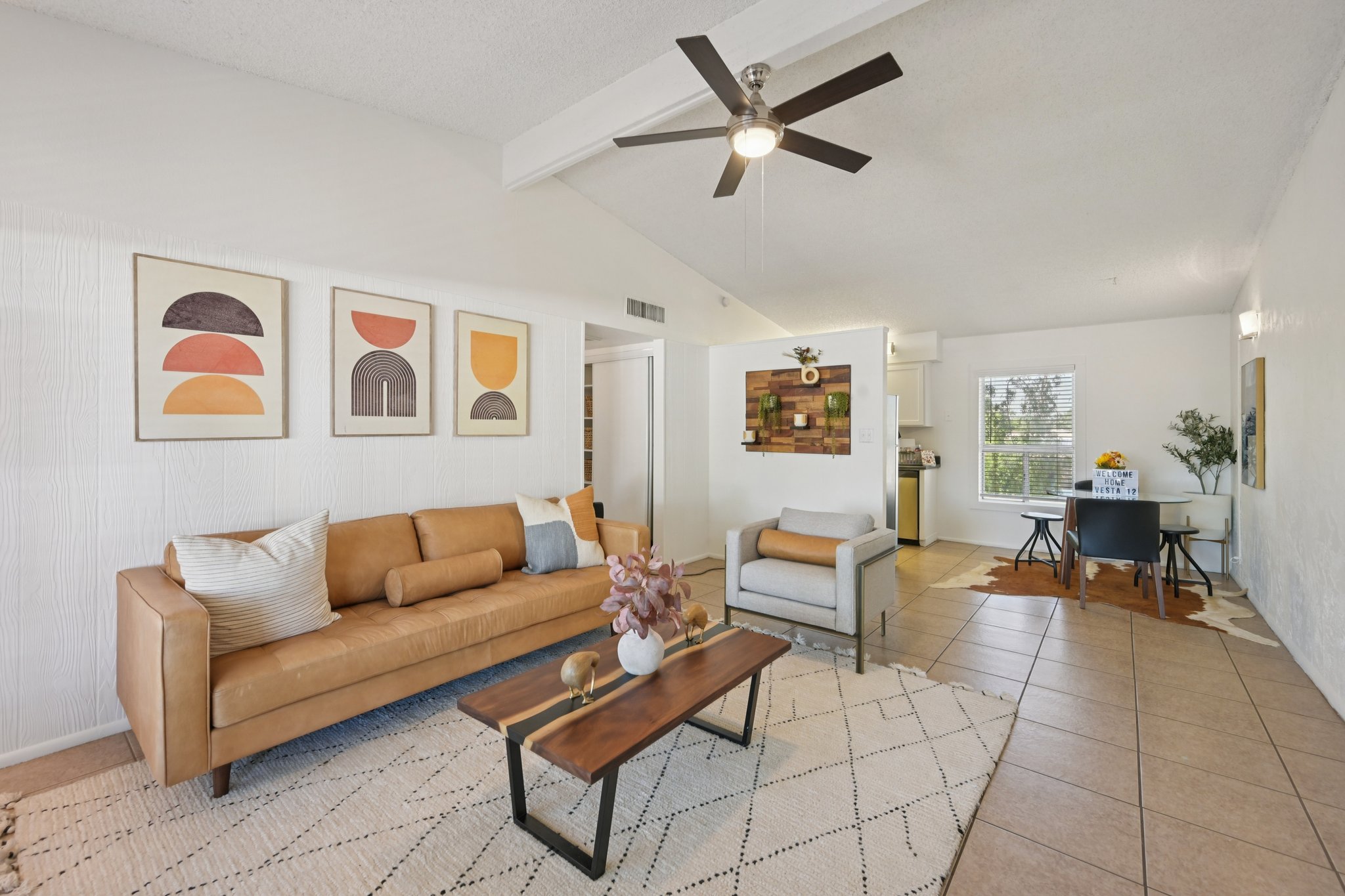 Modern living room featuring a tan leather sofa with decorative pillows, a minimalist coffee table, and wall art displaying geometric shapes in warm colors. The space has a ceiling fan and tile flooring, with a dining area visible in the background. Natural light streams in through the windows, enhancing the inviting ambiance.