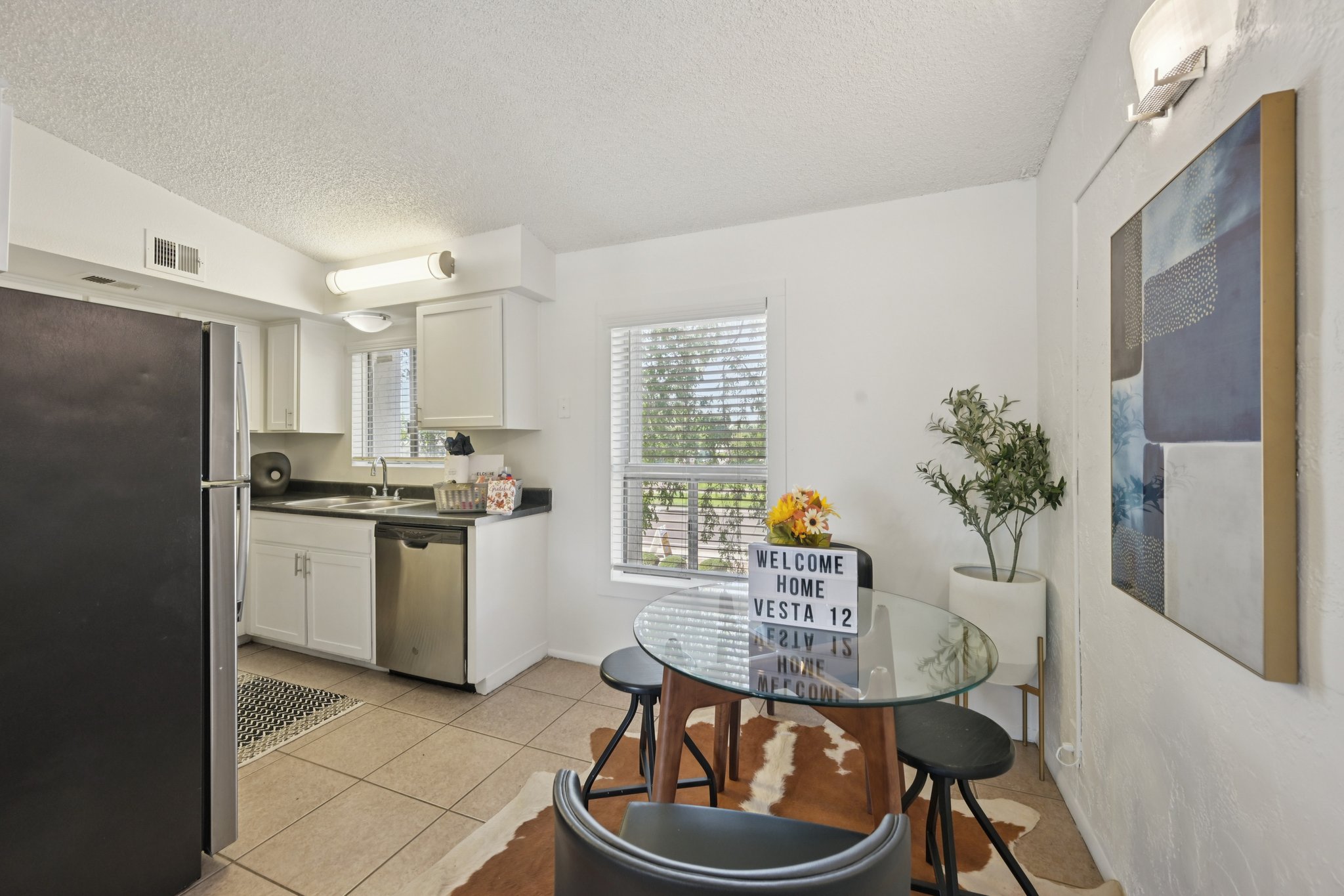 A bright, modern kitchen featuring a round glass dining table with black chairs, a small decorative plant, and a welcome sign. The kitchen has white cabinetry, stainless steel appliances, and a window with blinds, allowing natural light to illuminate the space. A cowhide rug adds warmth to the tile floor.