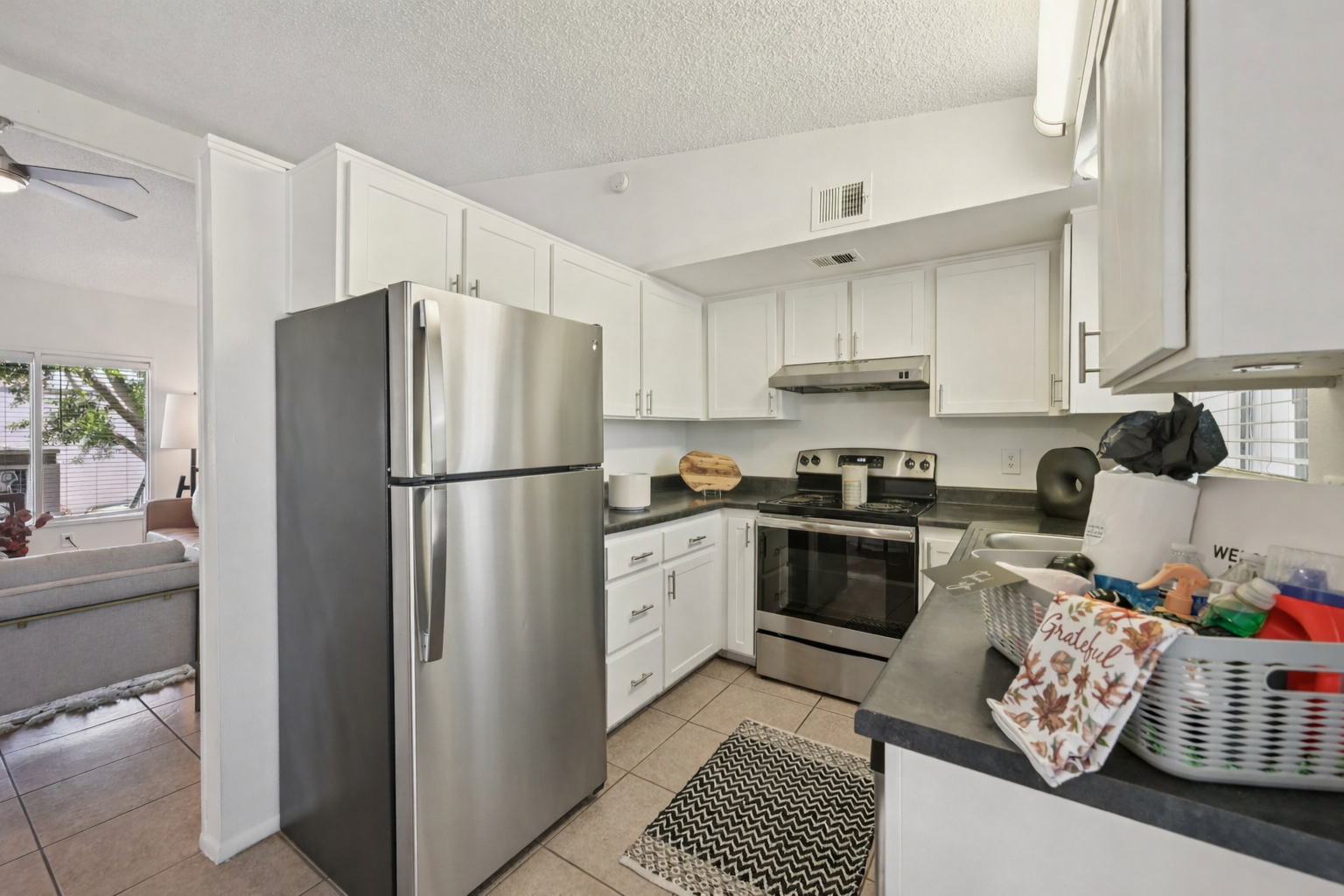 A modern kitchen featuring white cabinetry, stainless steel appliances, a black countertop, and a decorative rug. A bowl of fruit and various kitchen items are visible on the countertop, while a living area with a sofa is seen in the background through an open doorway.