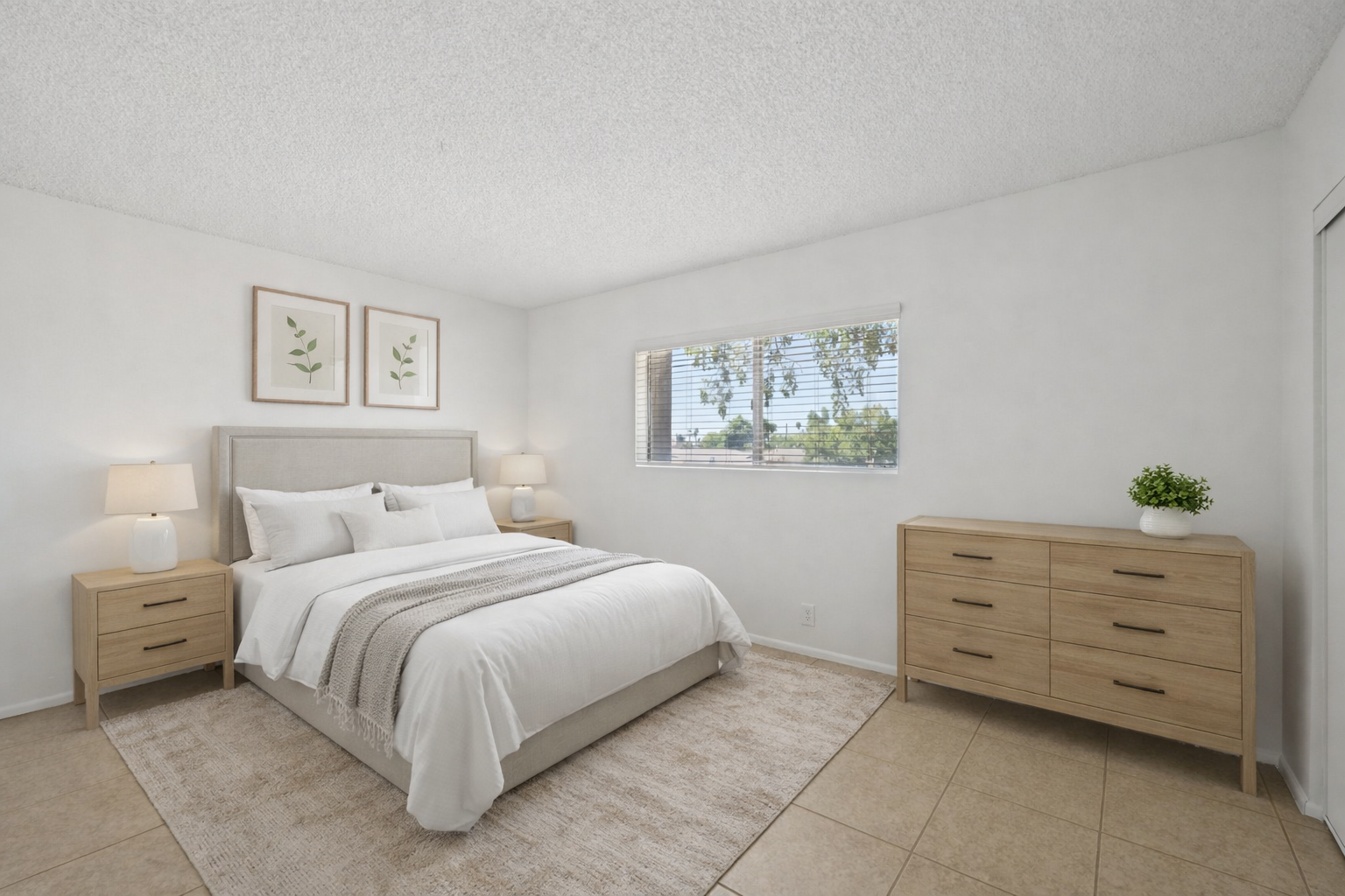A modern bedroom featuring a light color scheme. The room includes a large bed with white bedding, two bedside lamps, and framed botanical artwork on the walls. A window with natural light overlooks greenery outside, and there's a wooden dresser beside the bed, with a small plant on top.