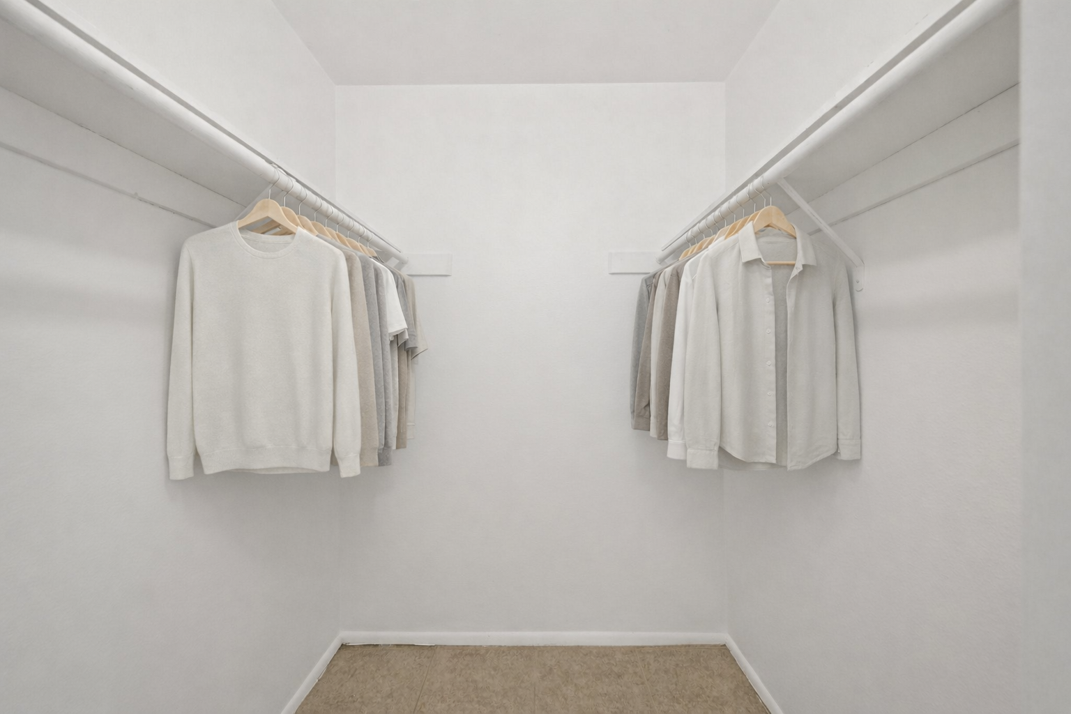 A minimalist walk-in closet featuring two rows of neatly hung clothing. The left side displays light-colored sweaters, while the right side has light-colored shirts. The walls and floor are plain white and beige, creating a clean and spacious appearance.