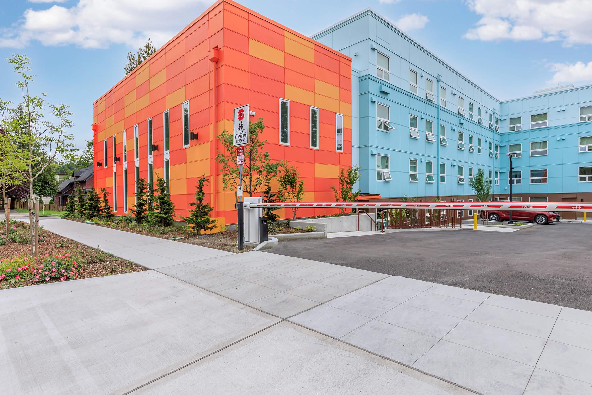Colorful modern building featuring orange and blue sections, surrounded by landscaped greenery and parking space. A barrier gate is visible in the entrance to the parking area. The scene is set against a clear blue sky with a few clouds.