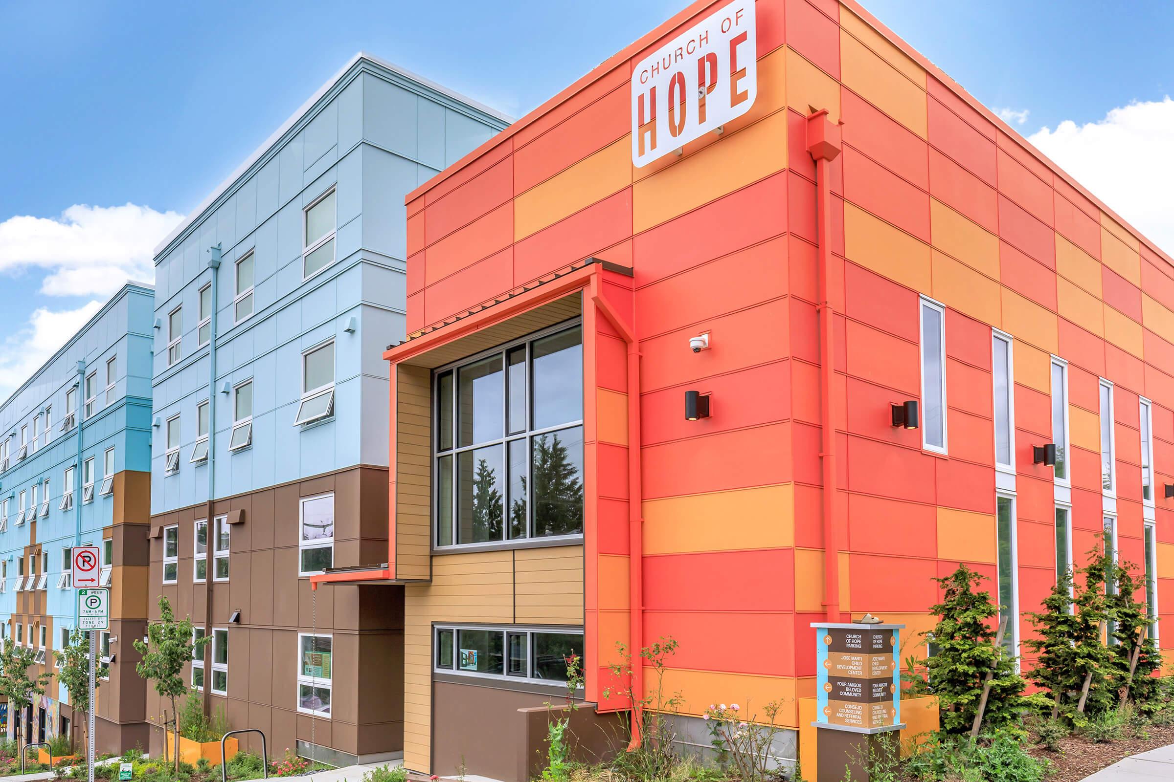 A modern building of the Church of Hope featuring a vibrant orange and red exterior, alongside a blue and brown structure. The entrance is clearly marked, and there are signs and greenery surrounding the area. The sky is clear with a few clouds.