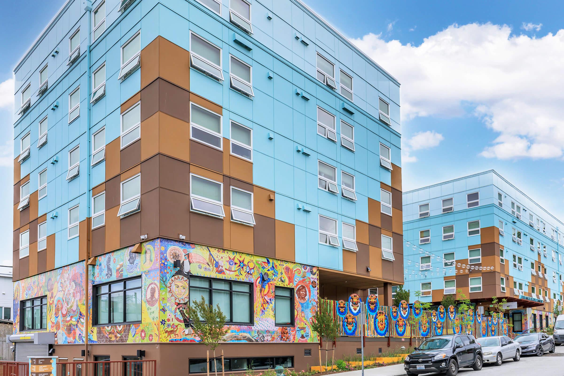 Colorful urban apartment buildings featuring a vibrant mural on the ground floor. The buildings have a modern design with blue and brown facades. The mural displays various artistic elements, adding a lively touch to the exterior. Several parked cars are visible in the foreground, with a clear blue sky above.