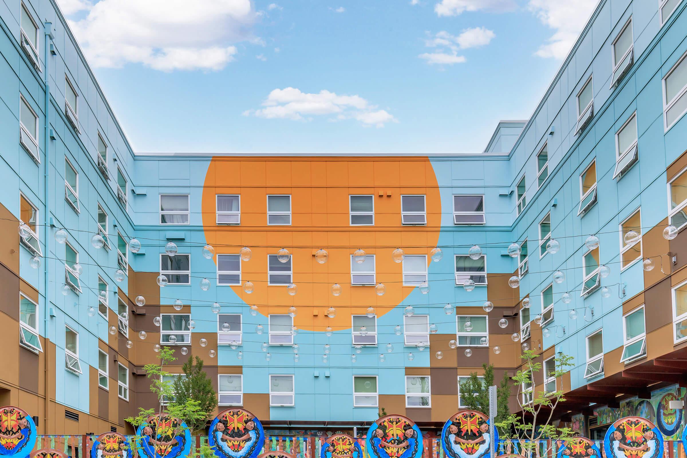A colorful modern building with a large orange circle on the front, surrounded by blue and brown panels. The courtyard features hanging light fixtures and circular designs on the ground, with greenery and a bright sky overhead.