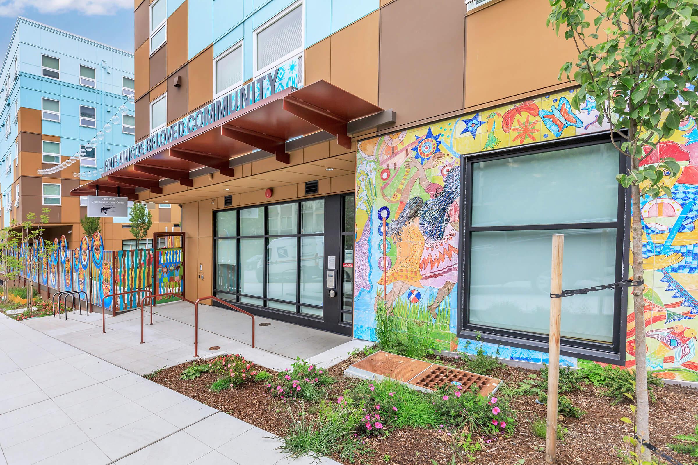 Vibrant community building entrance featuring colorful murals on the walls, with decorative landscaping and a welcoming atmosphere. The building has modern architecture, large windows, and a sign indicating it is a community space.