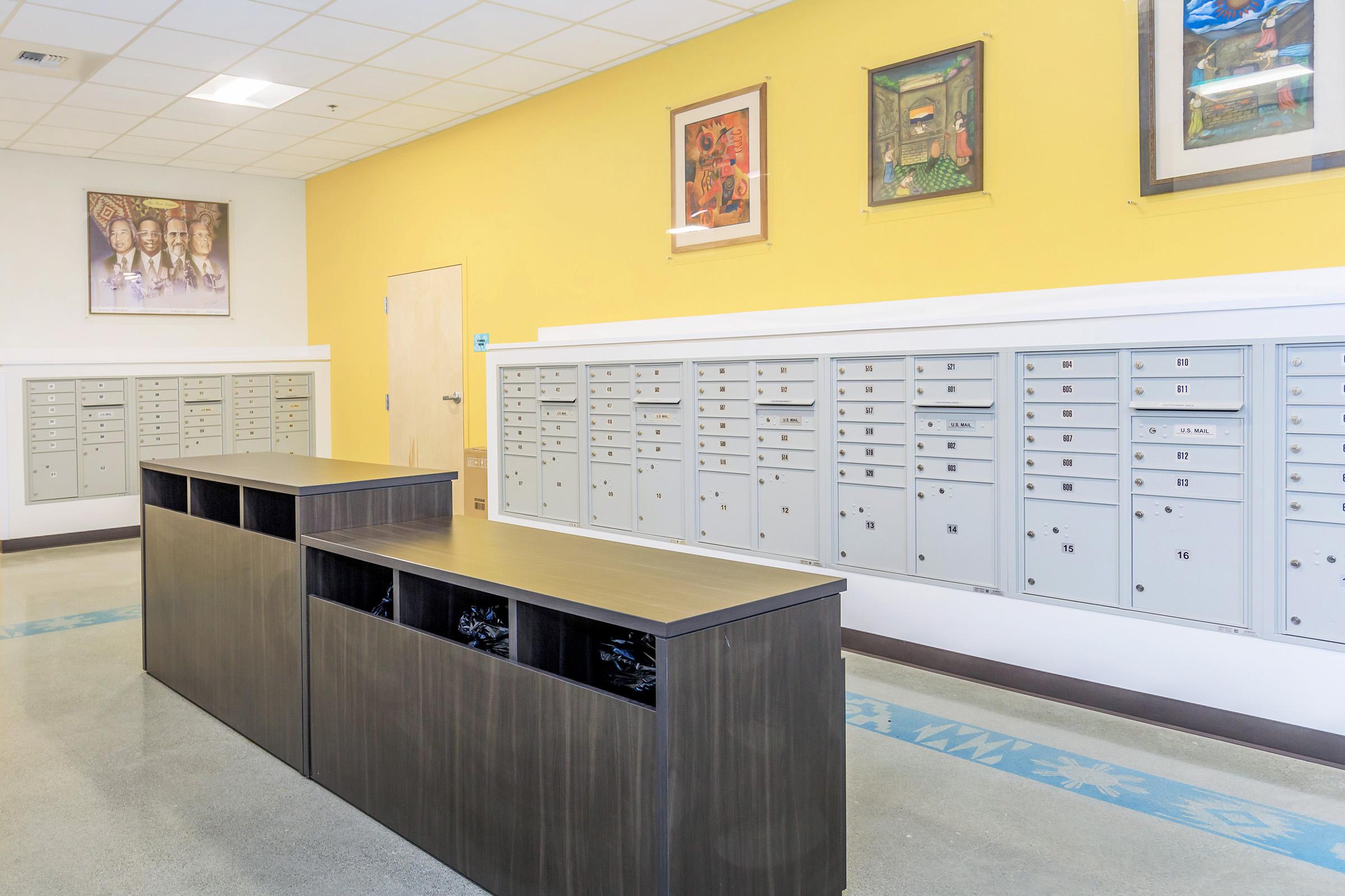 A well-lit common area featuring organized postal mailboxes along a wall with a vibrant yellow background. Two wooden tables are placed in the center, providing space for mail handling. The wall also has framed artwork, adding a decorative touch to the environment.