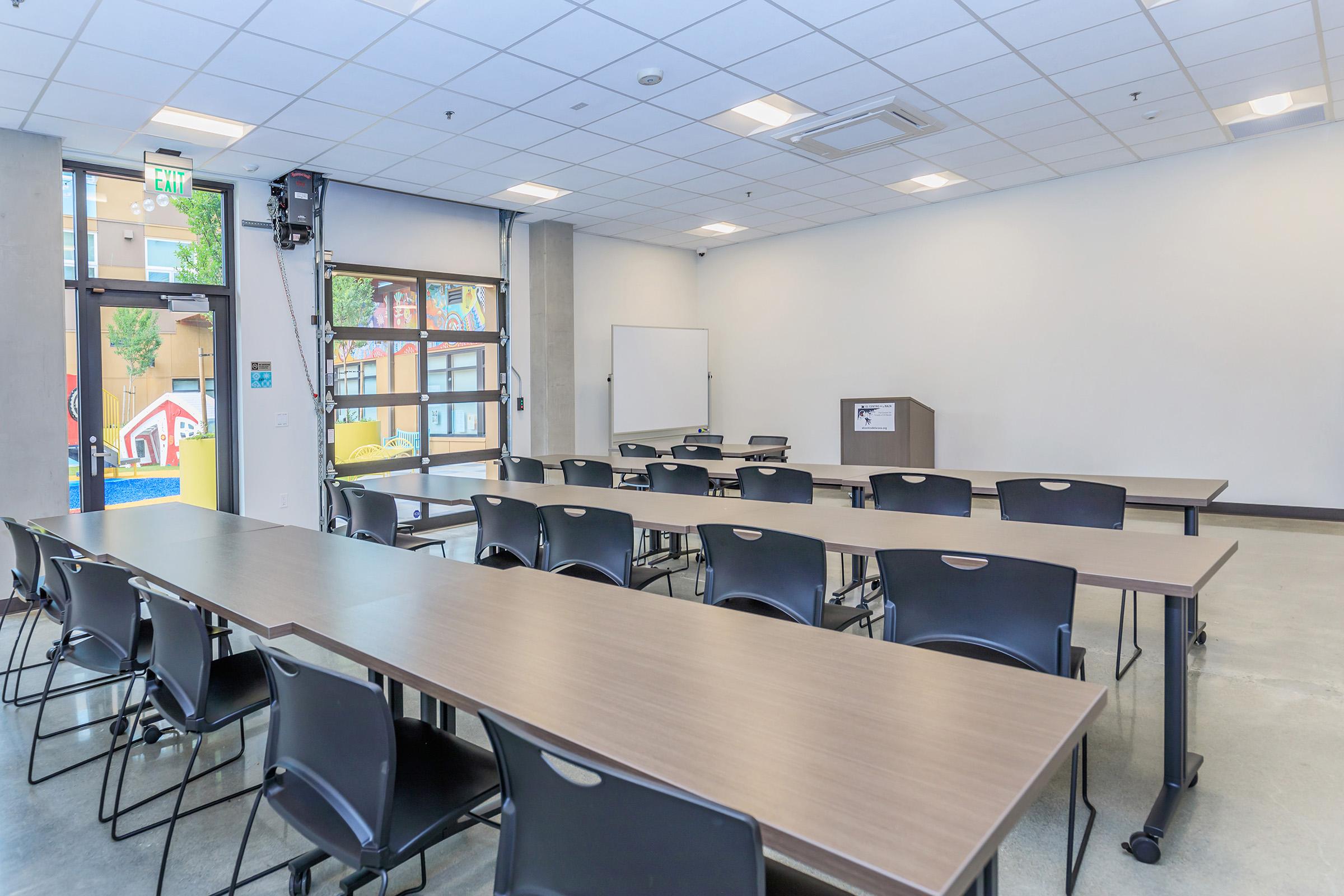 A modern classroom setup featuring multiple long tables arranged in a row with black chairs. The spacious room has large windows allowing natural light, a blank presentation board on the wall, and colorful artwork visible outside, creating an inviting learning environment.
