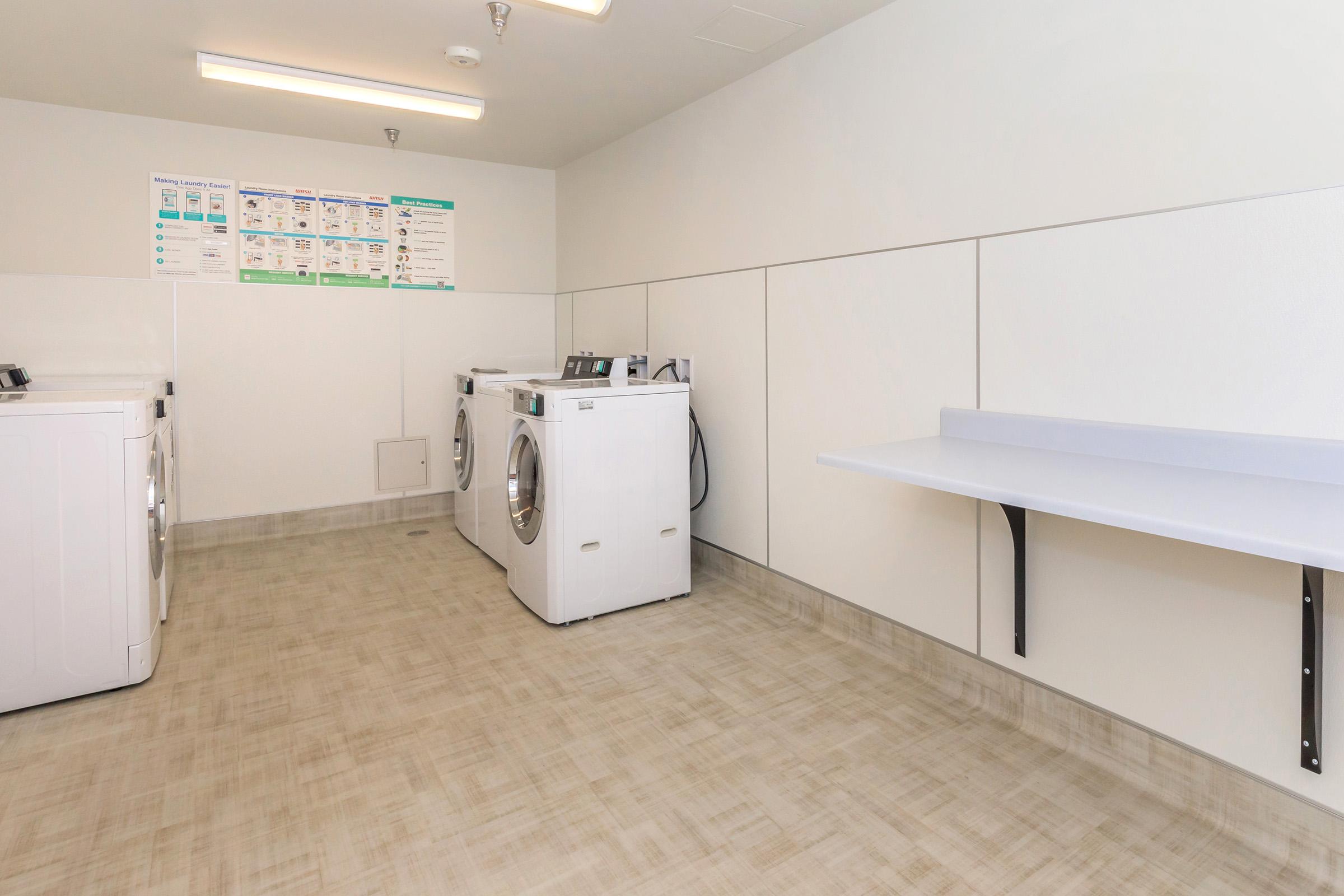 A clean and modern laundry room featuring several white washing machines against a plain wall. There is a countertop on the right side for folding clothes, and instructional signage on the wall above the machines. The floor is light-colored with a contemporary design.