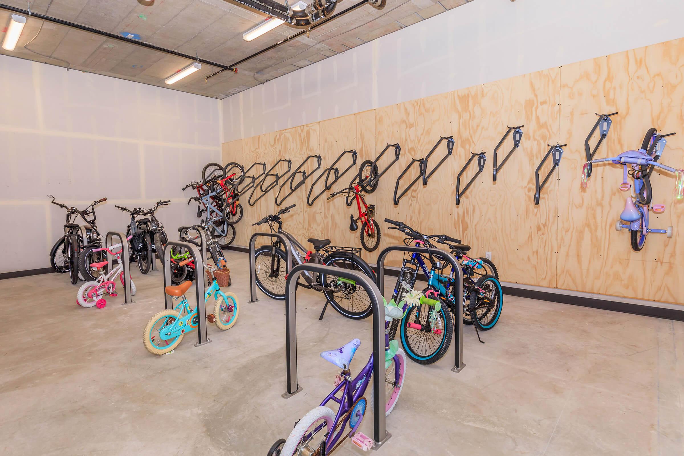 A well-organized bicycle storage room featuring various bikes neatly hung on wall-mounted racks. The floor is concrete, and the walls are unfinished with wooden panels. A mix of adult and children's bicycles, including brightly colored ones, is displayed in the space. Adequate lighting enhances visibility.