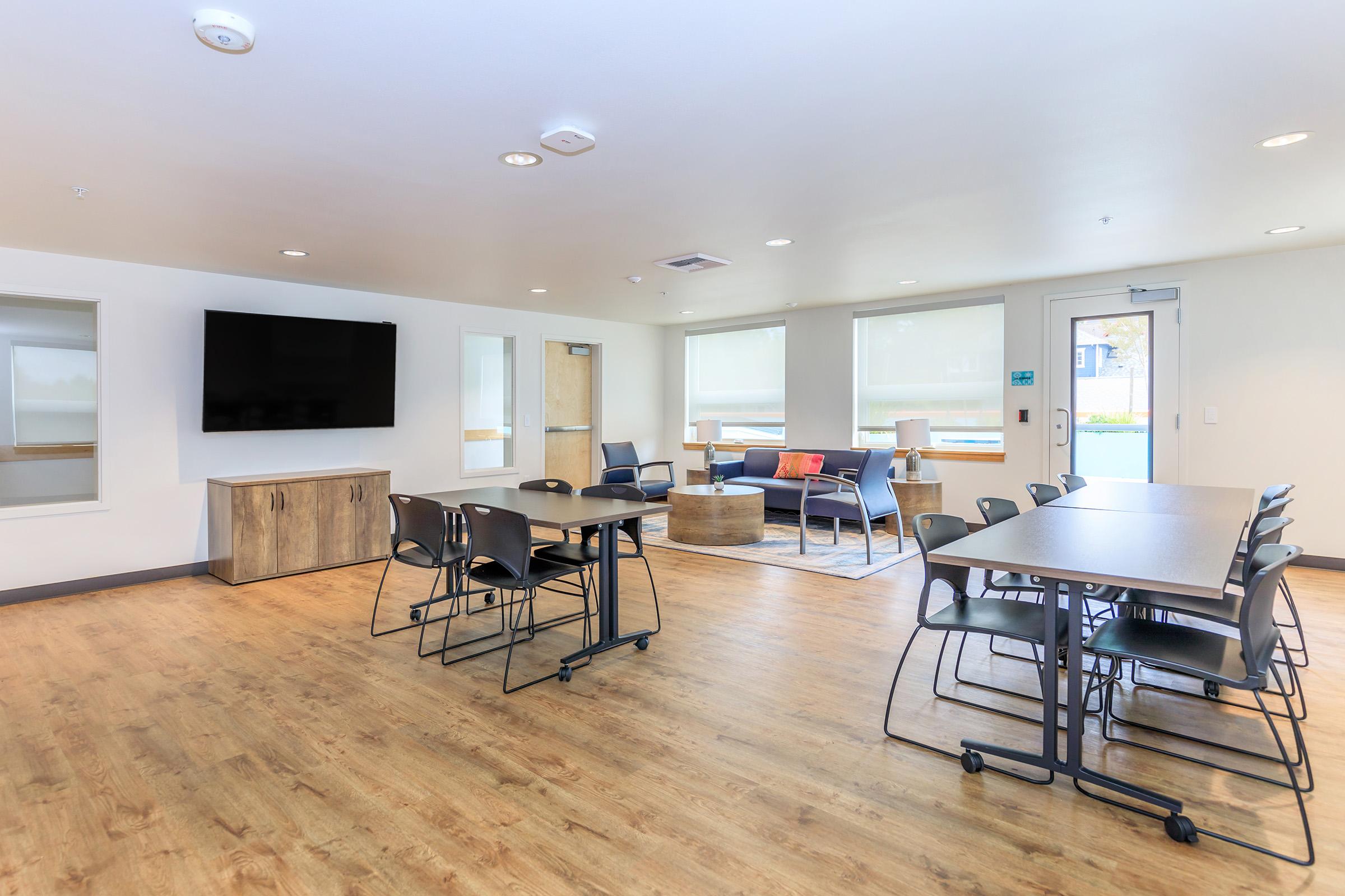 A spacious, modern common room featuring a large wall-mounted TV, a comfortable seating area with sofas and a coffee table, and multiple tables and chairs arranged for group activities. Large windows allow natural light to fill the space, which has a neutral color palette.