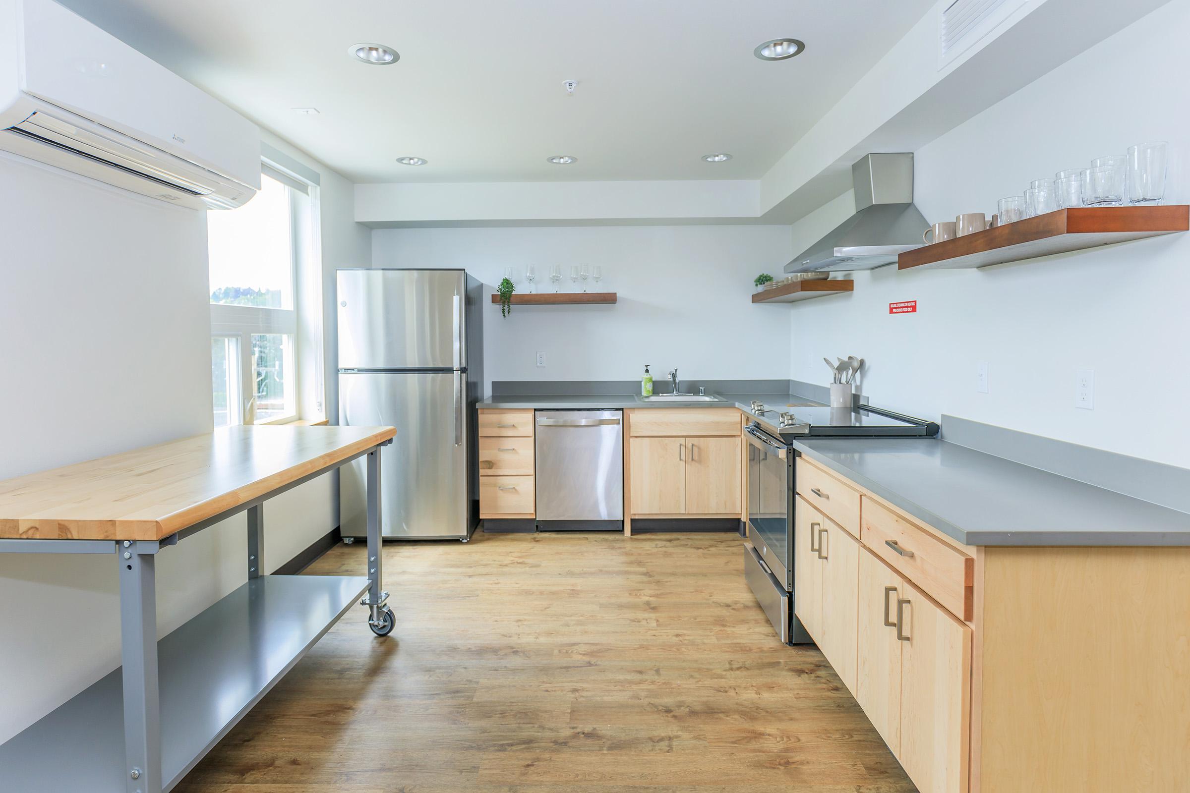Modern kitchen with stainless steel appliances, including a refrigerator, dishwasher, and stove. The countertops are gray with wooden cabinetry. Open shelving displays glasses. A large window allows natural light, and there's a wooden prep table on wheels for added workspace. Wood flooring completes the design.