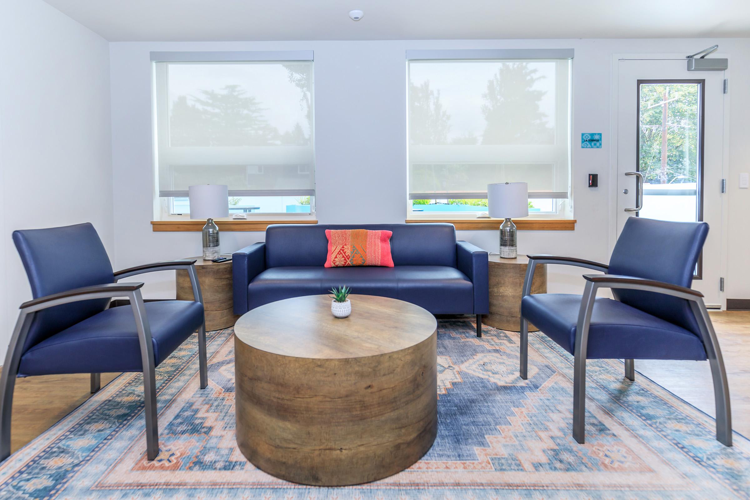 A modern seating area featuring two blue armchairs and a navy sofa with a colorful pillow. A round wooden coffee table sits in the center, adorned with a small plant. Two lamps are positioned on either side of the sofa, and large windows allow natural light into the space.