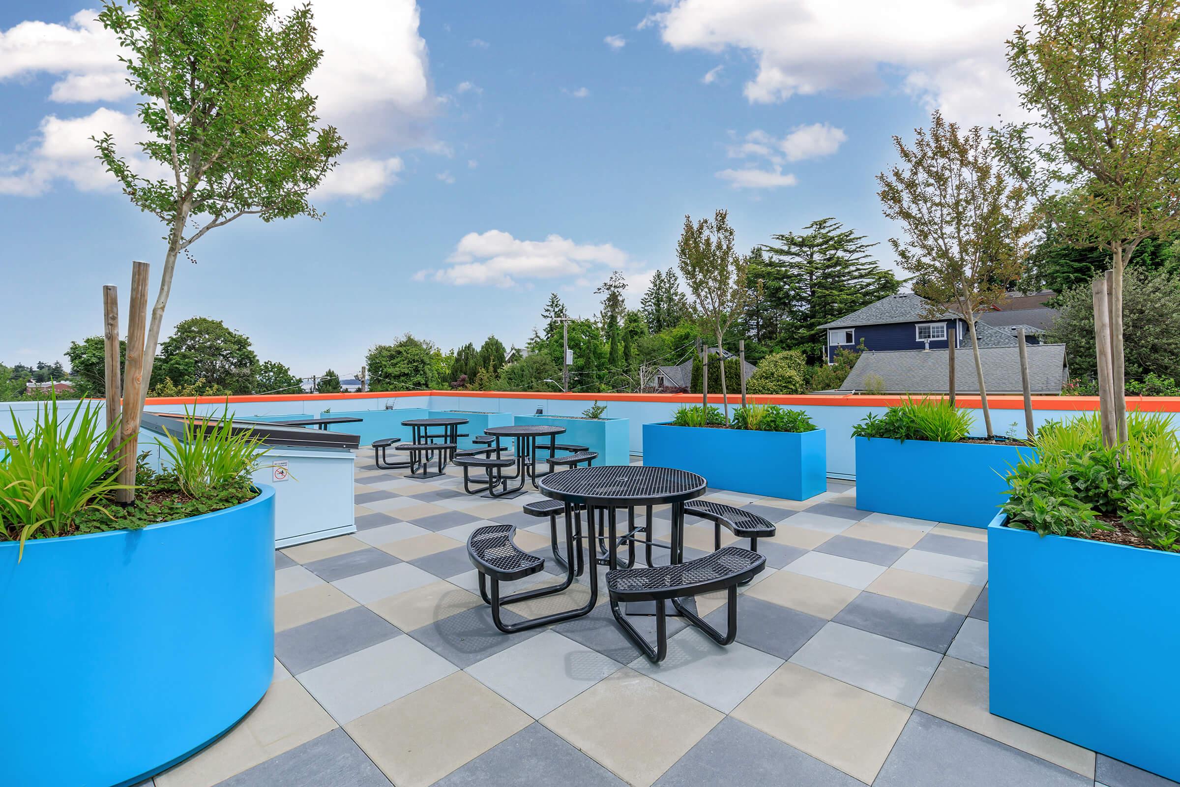 A rooftop patio featuring several black tables and chairs surrounded by large blue planters filled with greenery. The area is paved with gray tiles and is set against a backdrop of trees and houses under a clear blue sky with soft clouds.