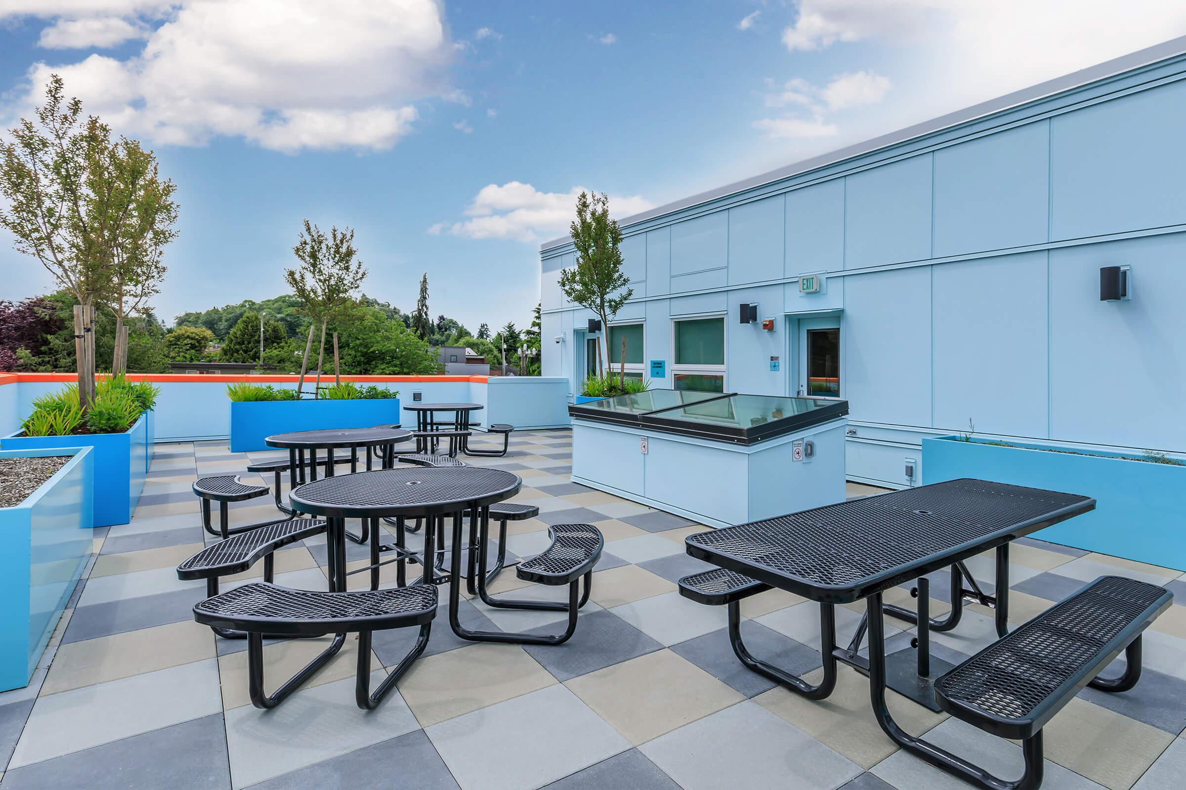 Rooftop terrace with multiple black picnic tables surrounded by blue planters and trees, set against a backdrop of a clear sky and greenery. The space is designed for relaxation and social gatherings.