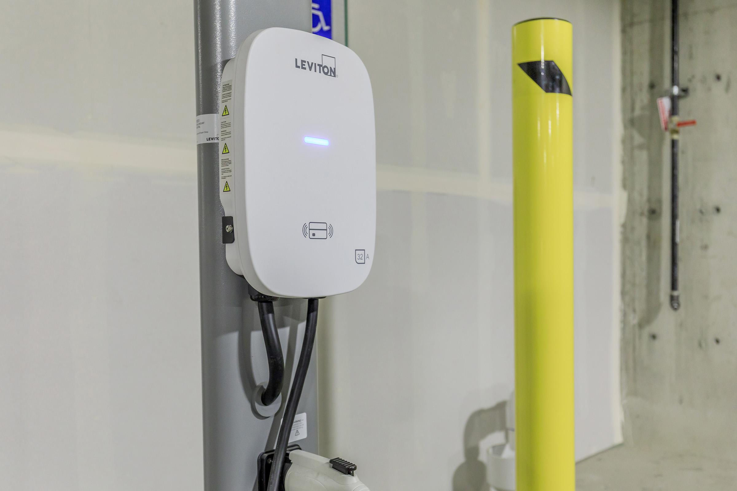A Leviton electric vehicle charging station mounted on a wall, featuring indicator lights and connected cables. A yellow pole stands nearby, indicating the parking area for electric vehicles. The background shows an unfinished concrete wall typical of a garage or parking structure.
