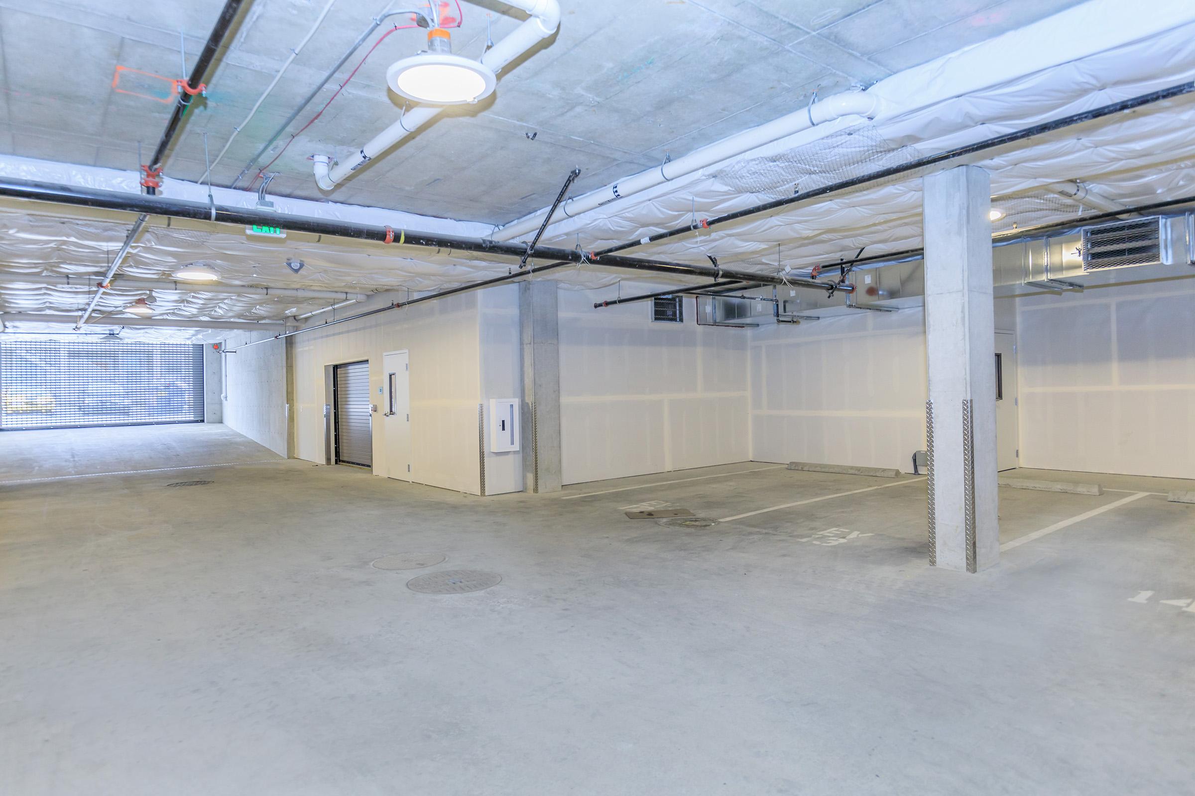 A clean, spacious underground parking garage featuring concrete walls and floors, overhead lighting, and several empty parking spaces. The entrance is visible in the background, and there is a roll-up door on the left side. Ductwork and pipes are installed along the ceiling.