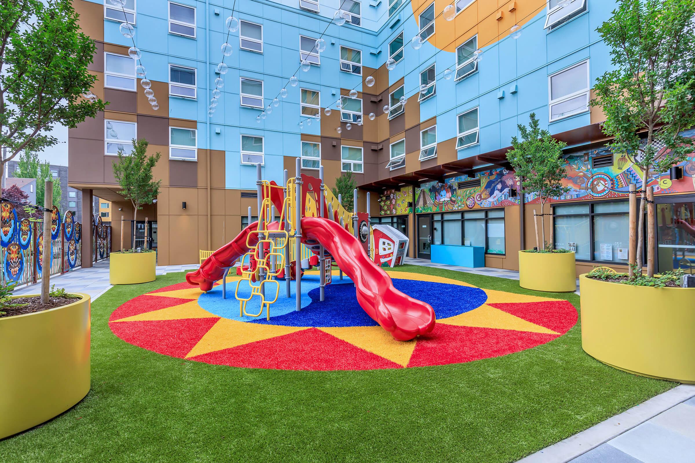 Vibrant outdoor playground featuring two red slides, surrounded by colorful soft flooring in a circular design. The area is enclosed by modern buildings with large windows and decorated walls, along with planters filled with small trees, creating a playful and inviting atmosphere for children.