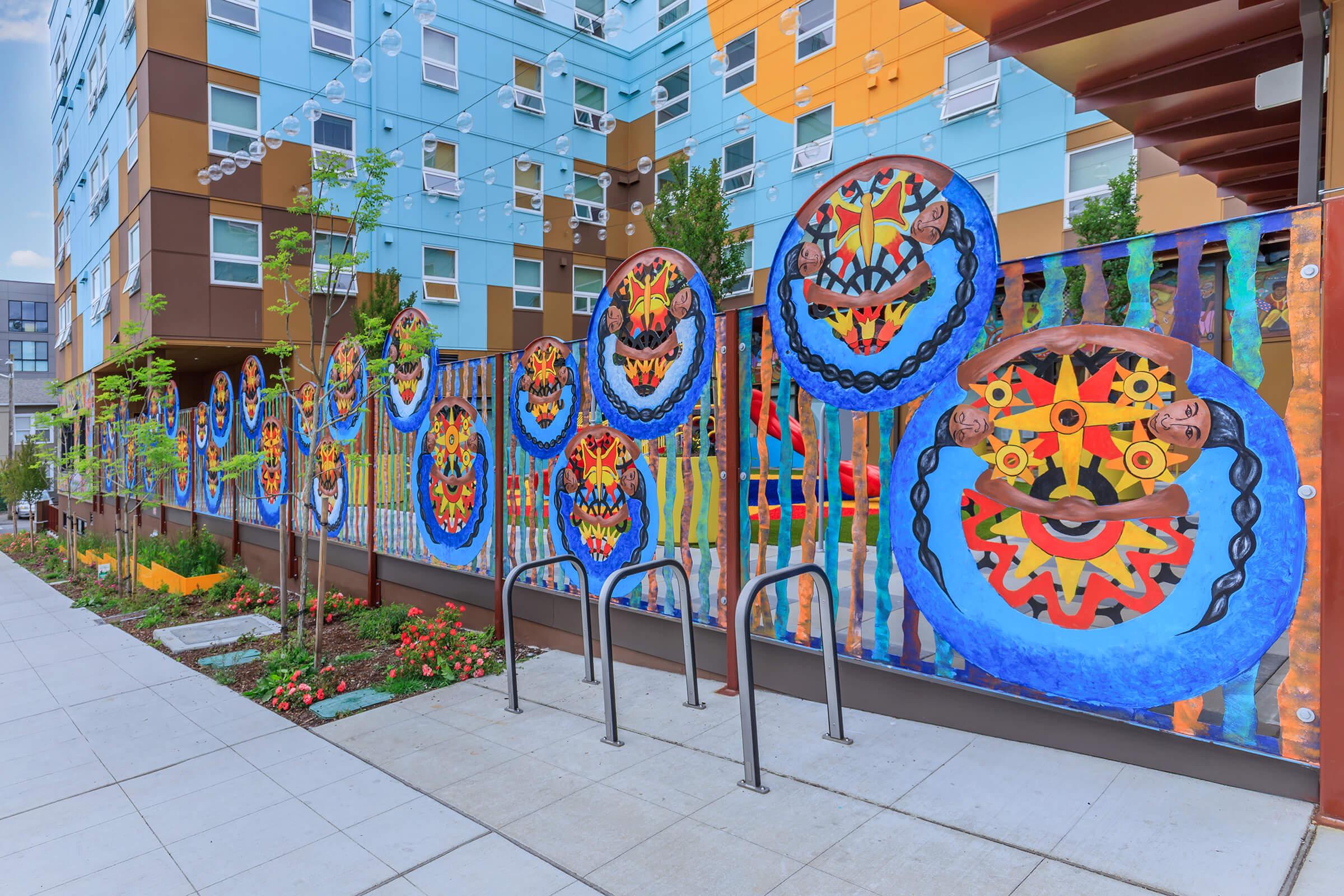 A colorful mural featuring circular designs with faces and abstract patterns, displayed on a fence next to a pathway. The background includes modern apartment buildings in various colors, with greenery and landscaping elements like flowers and bike racks visible.