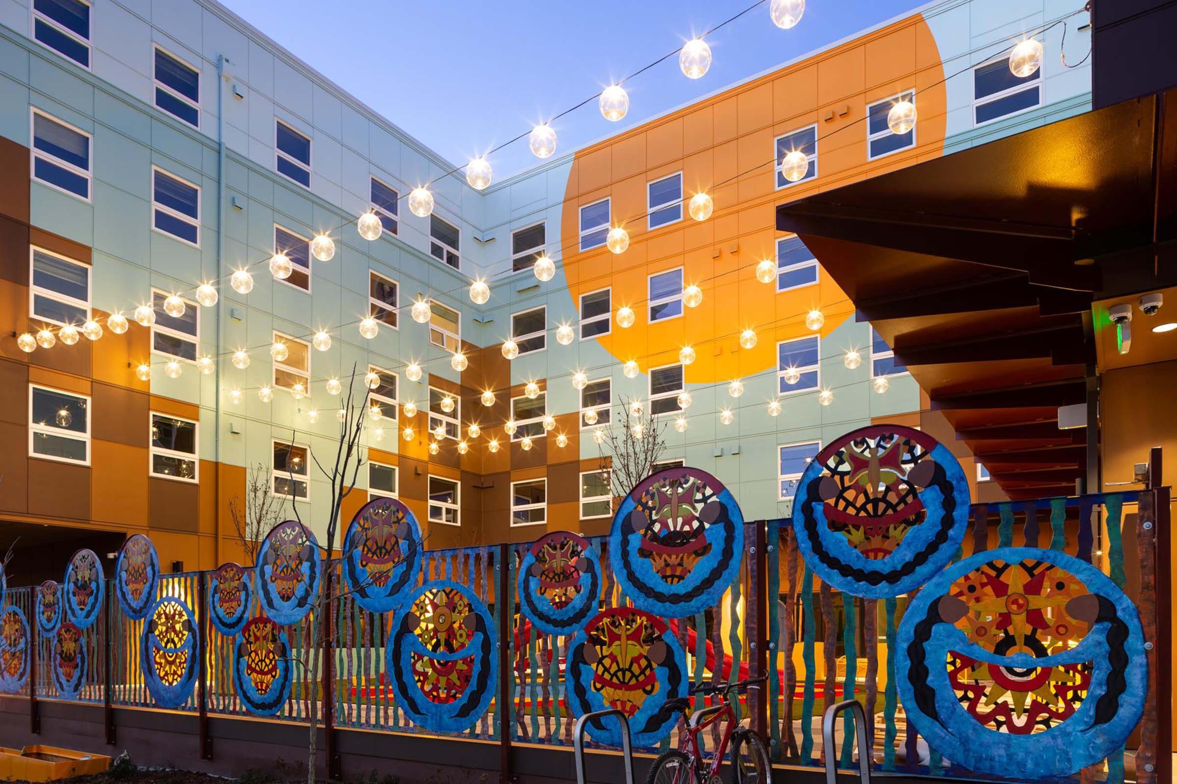 Colorful building exterior with vibrant murals and decorative circular art pieces hanging along a fence. Warm string lights illuminate the courtyard area at dusk, creating a lively atmosphere. The architecture features a mix of colors, including blues, oranges, and yellows, contributing to a welcoming environment.