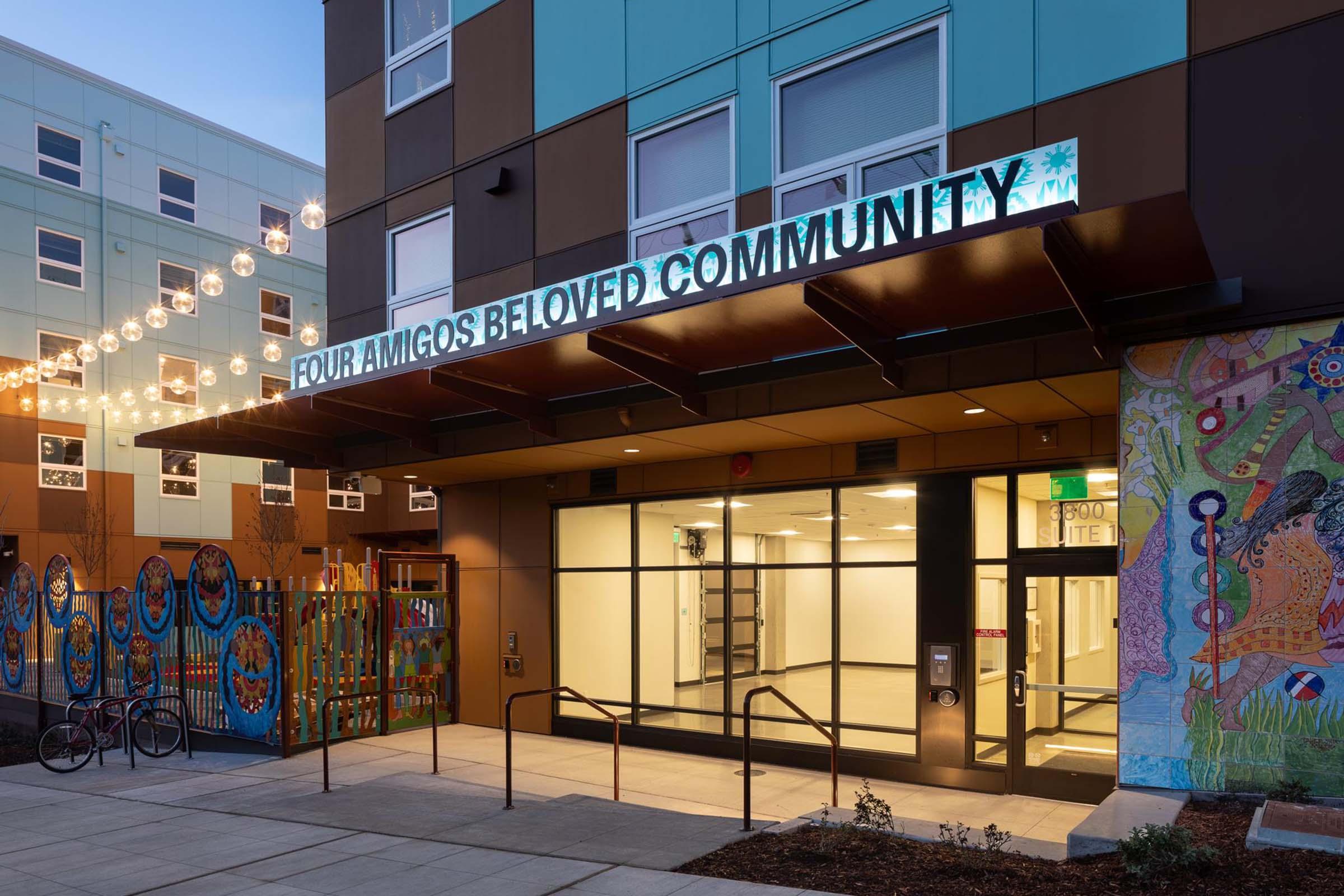 Entrance of the "Four Amigos Beloved Community" building, featuring a colorful facade with illuminated signage. The area is decorated with vibrant murals, and there are decorative lights strung above. The entrance has glass doors and accessible steps, and a bike rack is visible nearby.